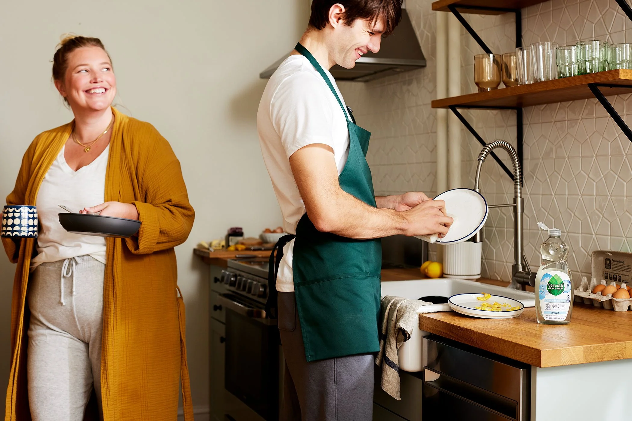 A young man and woman preparing breakfast in a modern kitchen. The man is washing dishes at the sink, while the woman, holding a coffee mug and a plate, smiles and watches him.