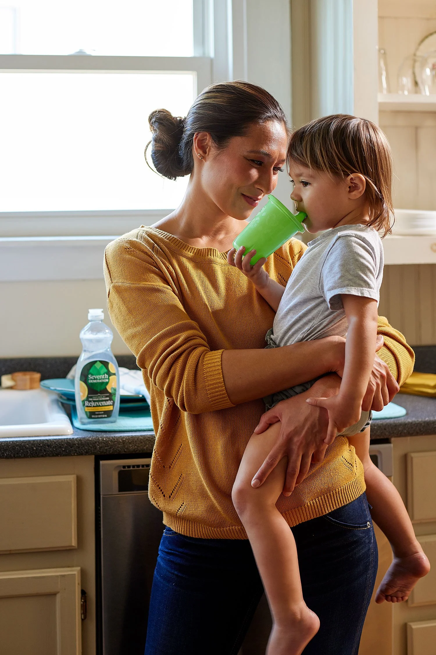 A woman holding a young boy in a kitchen, giving him a drink from a green cup. The woman has dark hair tied back, and the boy has light brown hair. There is a bottle of hand sanitizer on the counter behind them.