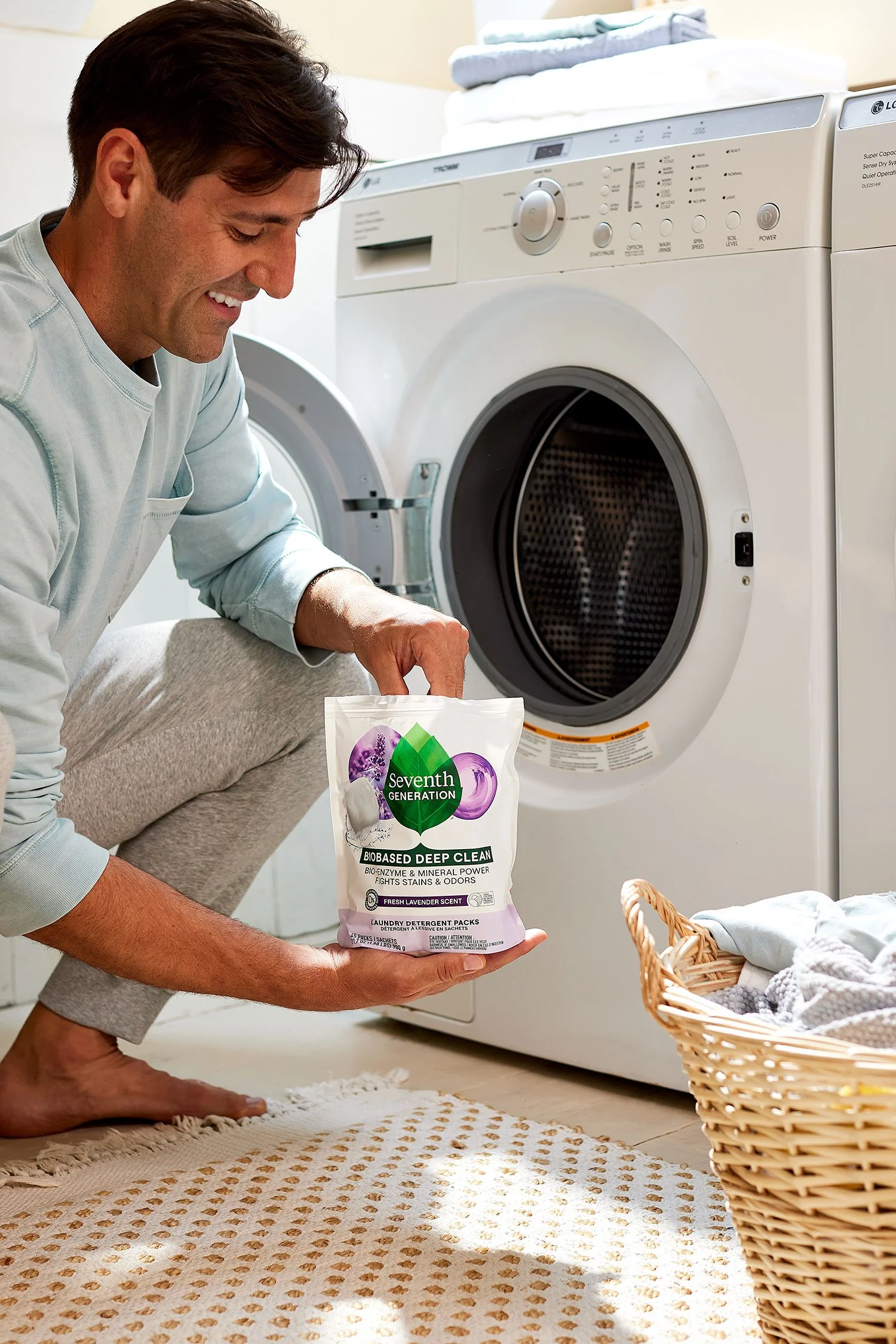 A man is kneeling on a beige rug holding a box of Seventh Generation laundry detergent in front of a front-loading washing machine.