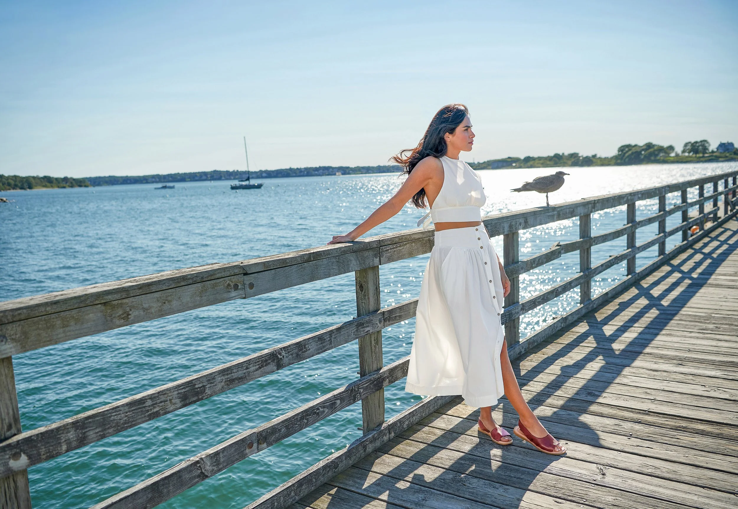 Woman in white outfit leaning on a wooden railing by a water body, with sailboats and a bird nearby on the railing, sunny sky.