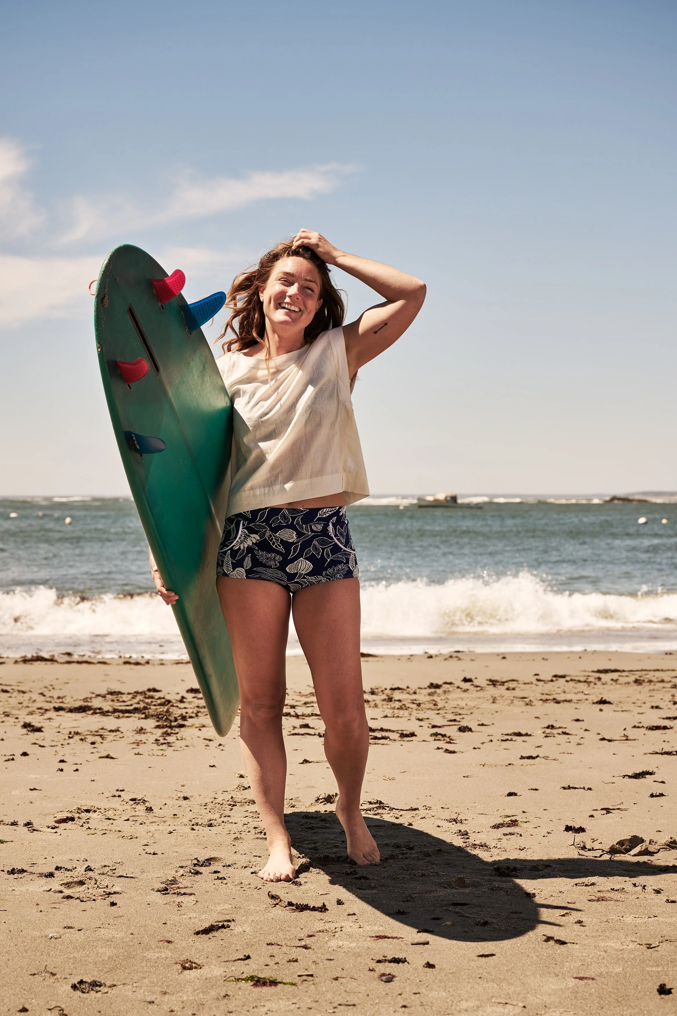 Woman standing on the beach holding a skateboard, smiling, with the ocean and sky in the background.