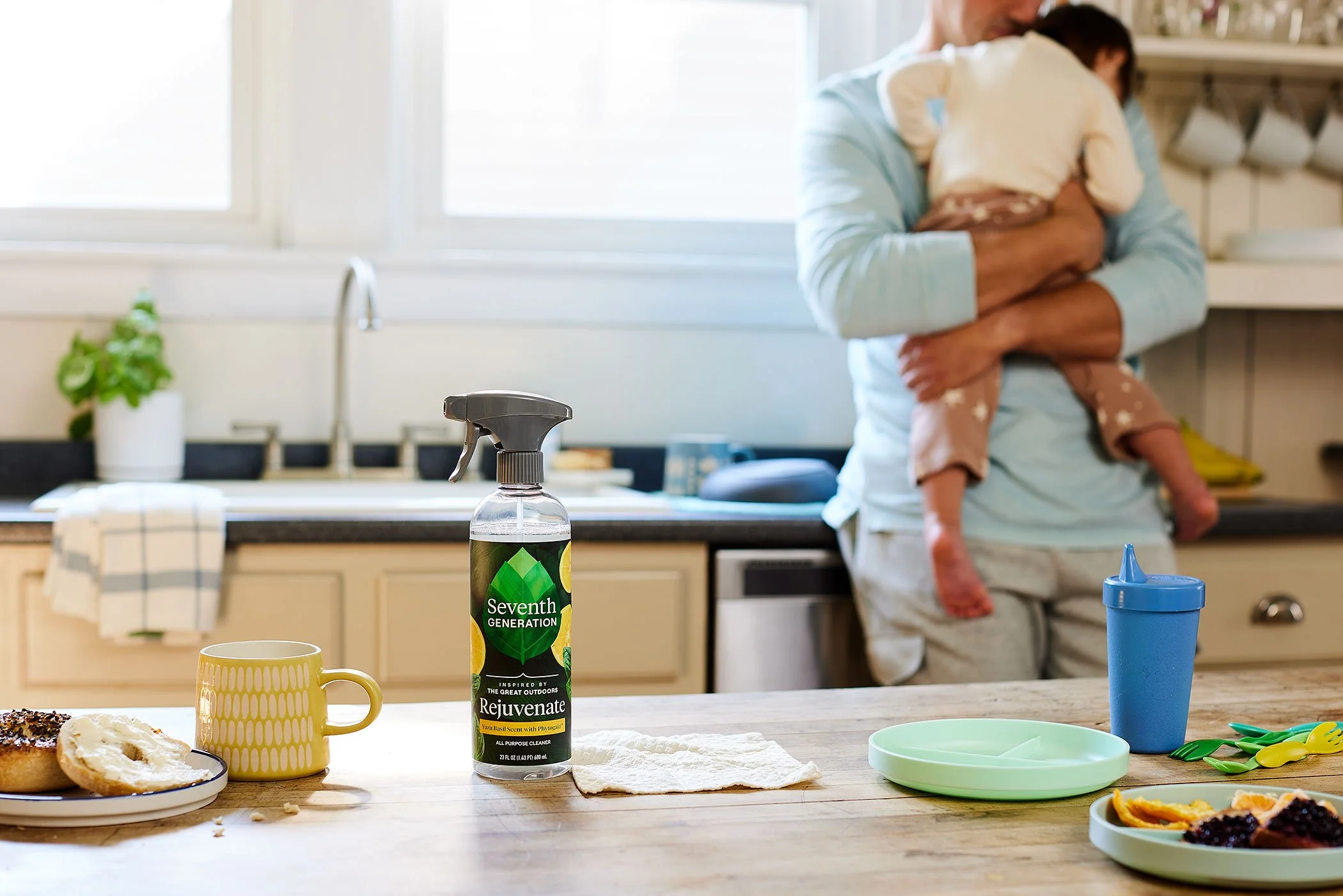 Kitchen counter with a spray bottle labeled 'Seventh Generation Rejuvenate', a yellow coffee mug, a green divided plate with food, a blue sippy cup, and utensils, with a person holding a child in the background near the sink.