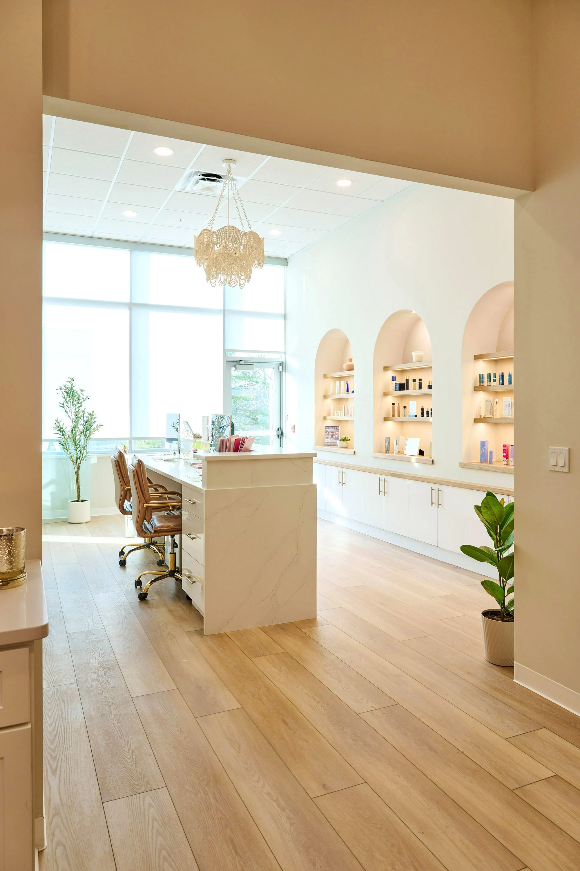 Modern, bright office or reception area with large windows, wooden flooring, white walls, a chandelier, a desk with chairs, and built-in shelves with decor and products.