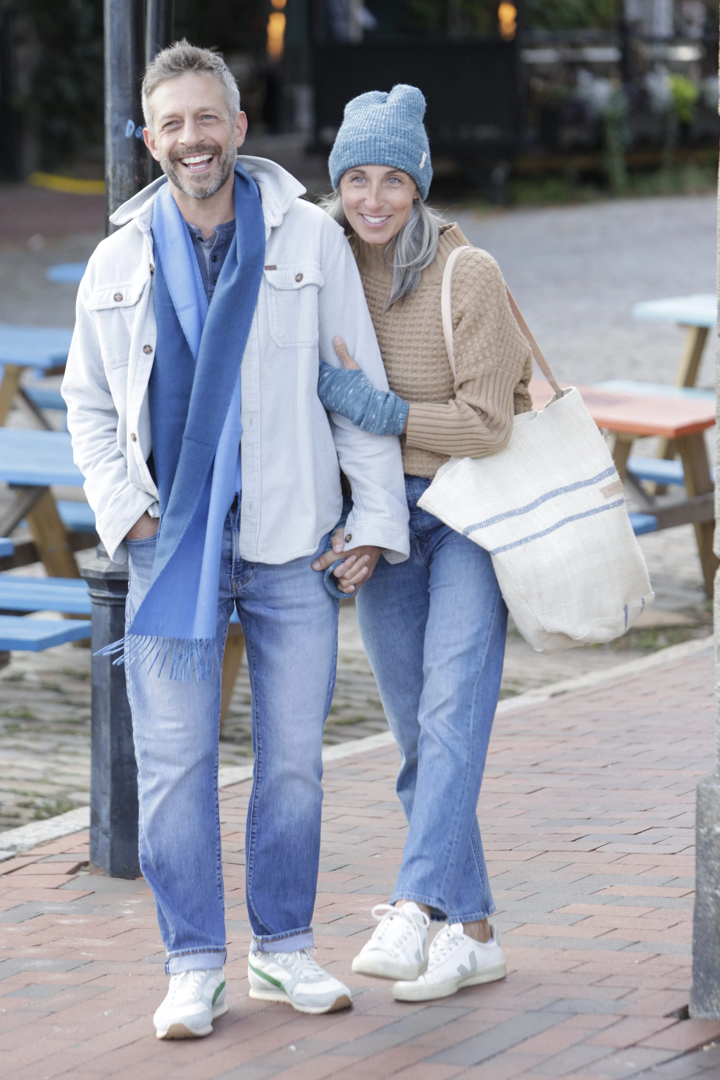 A smiling couple walking outdoors on a brick pathway, dressed casually in jeans, with the woman wearing a blue beanie and carrying a large tote bag.