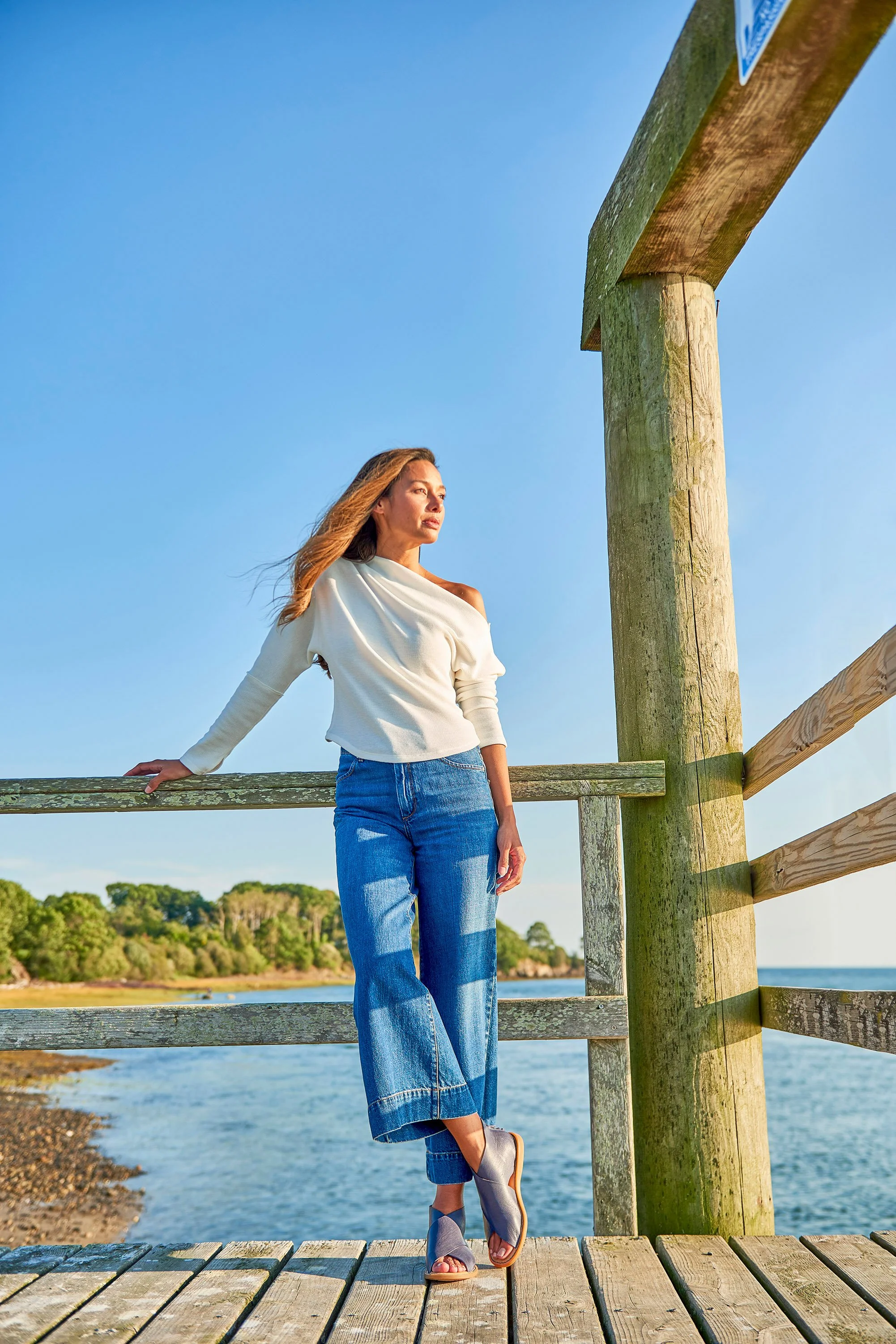 Woman standing on a wooden dock near the water on a sunny day with a blue sky and green trees in the background.