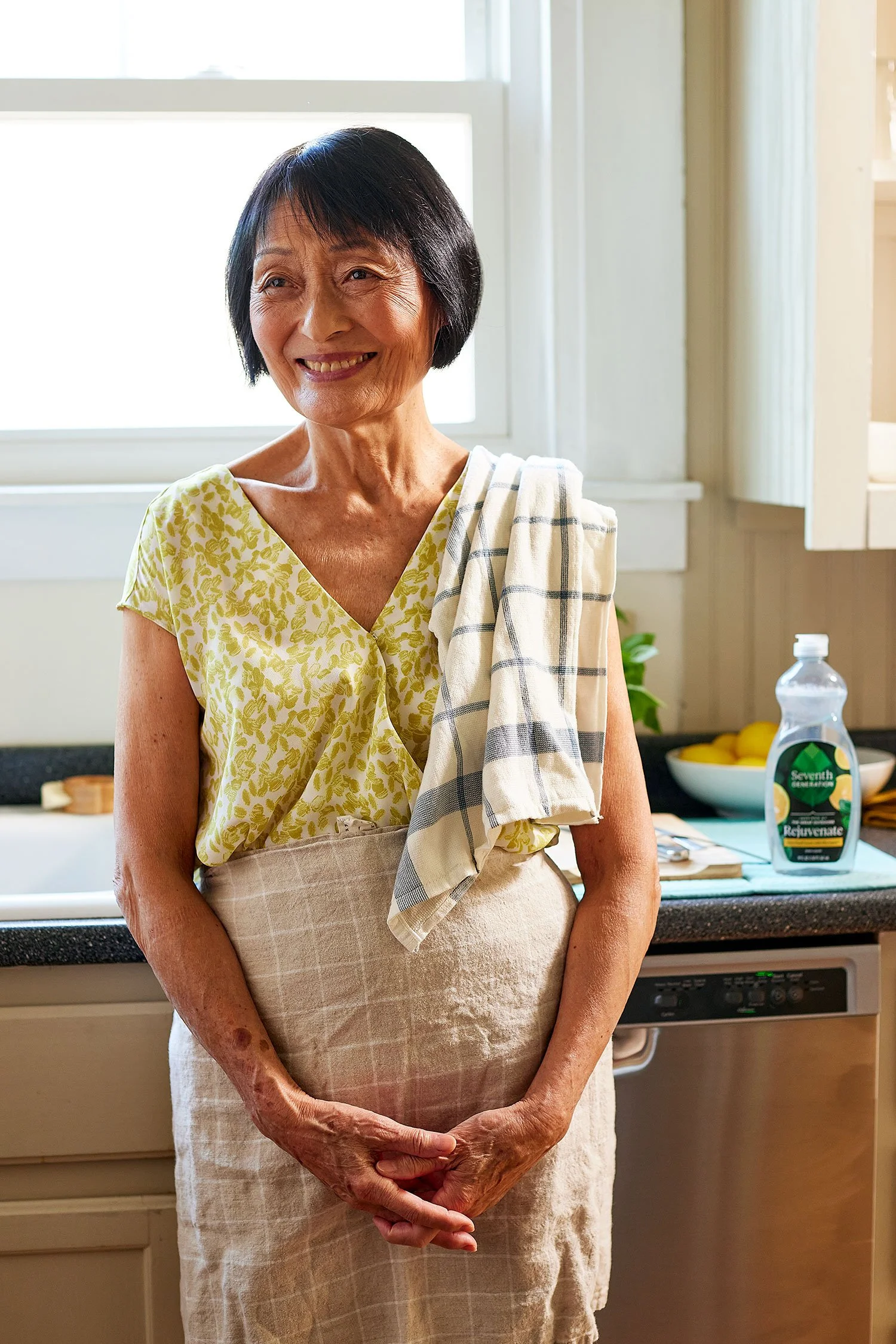 A smiling woman with short black hair stands in a kitchen, wearing a yellow patterned top and a beige apron, with a kitchen towel draped over her shoulder. The kitchen has a window, a bowl of lemons, and a bottle of beverage on the counter.