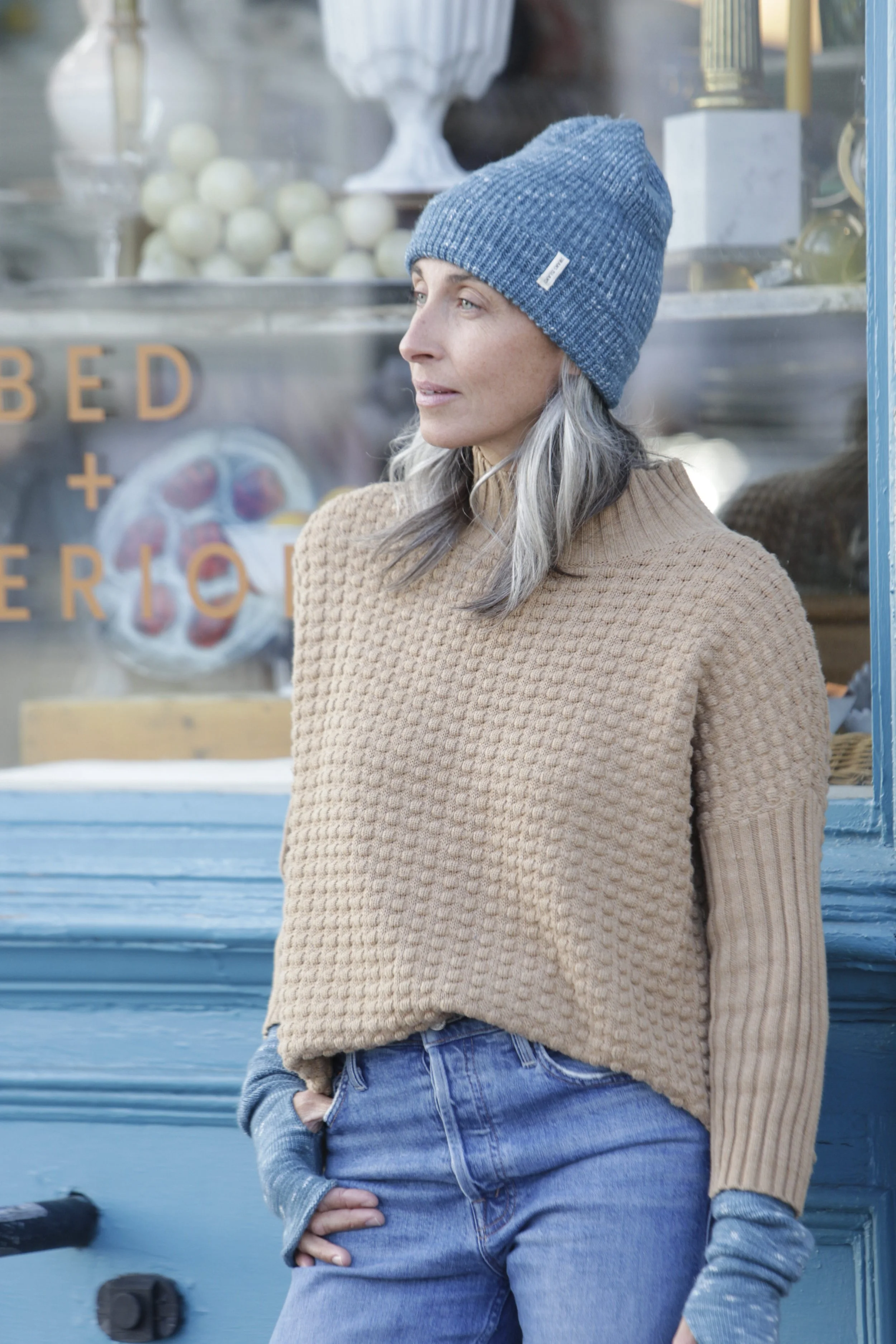 A woman with gray hair wearing a blue knit hat, beige sweater, and denim jeans standing outside a shop window.