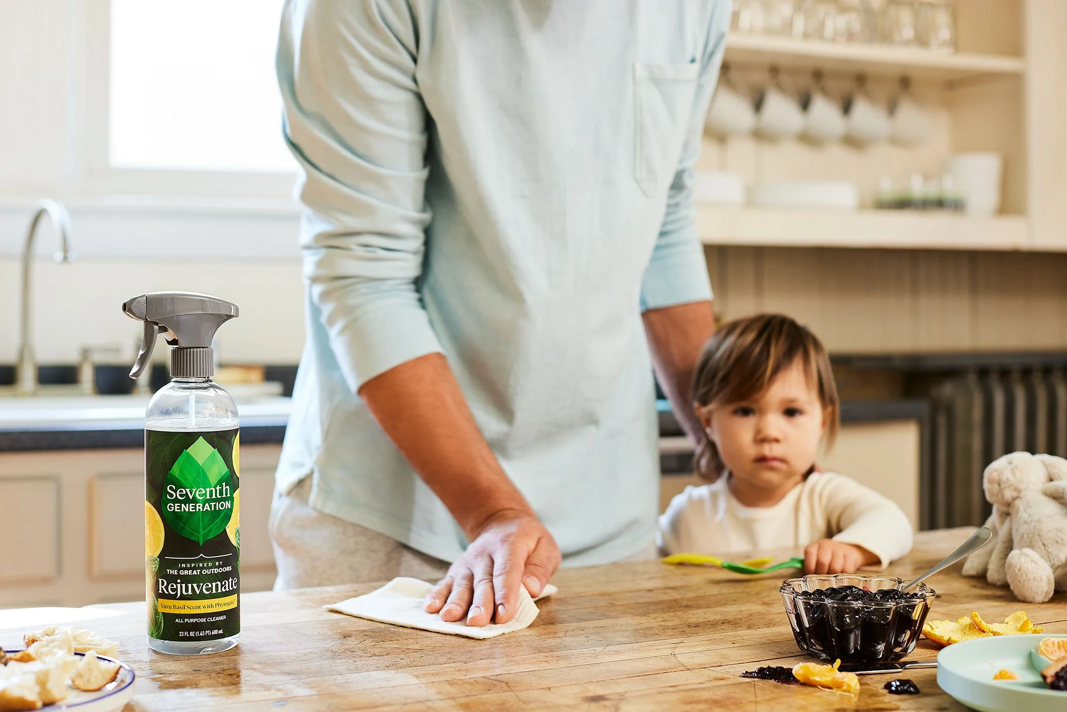 A man cleaning a wooden kitchen island with a cloth near a bottle of Seventh Generation all-purpose cleaner, with a young child sitting next to him and a bowl of dark berries on the table.