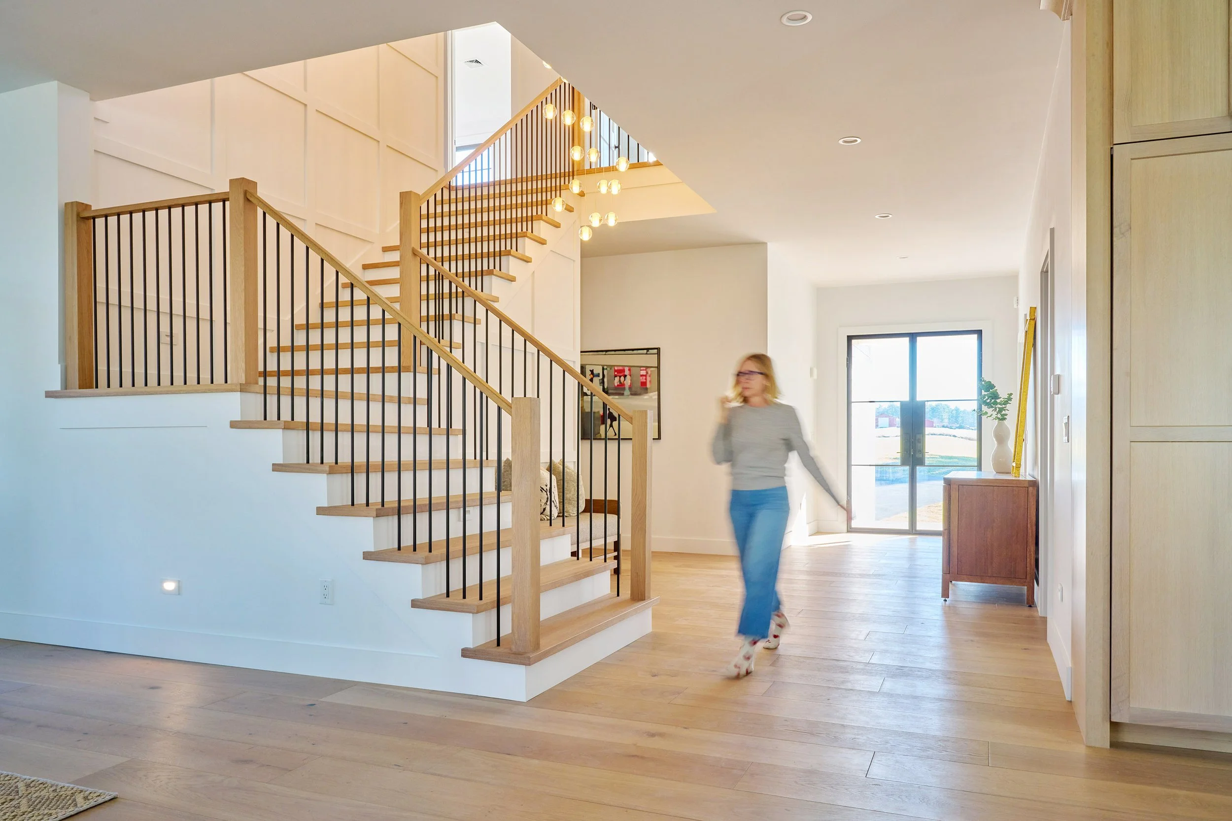 Interior of a modern home with wooden flooring, a staircase with wooden railings, a woman in a gray sweater and jeans walking and talking on her phone, a side table with a vase and green plant, large sliding glass door with outdoor view, and a bright, open living space.