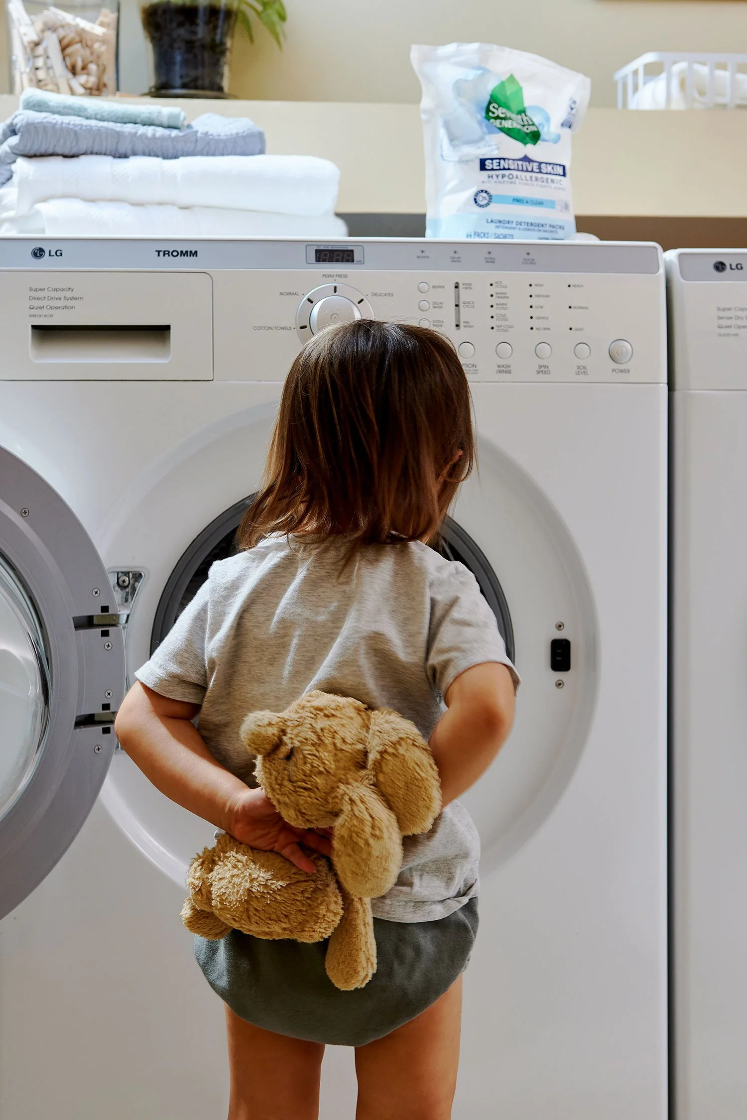 A young child with brown hair standing in front of a washing machine, holding a brown teddy bear behind their back. The child is wearing a gray t-shirt and dark shorts, in a laundry room with folded towels on top of the washing machine and detergent 