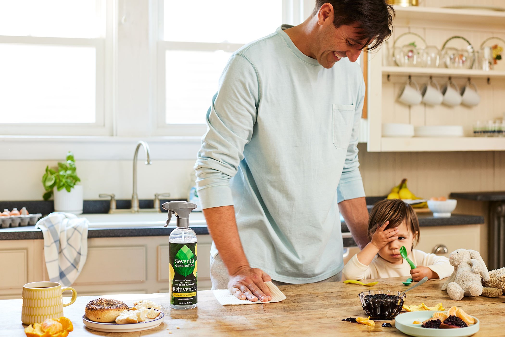 A man and a young child in a kitchen, with the man smiling and the child covering one eye with one hand, holding a spoon in the other, surrounded by breakfast foods and cleaning supplies.