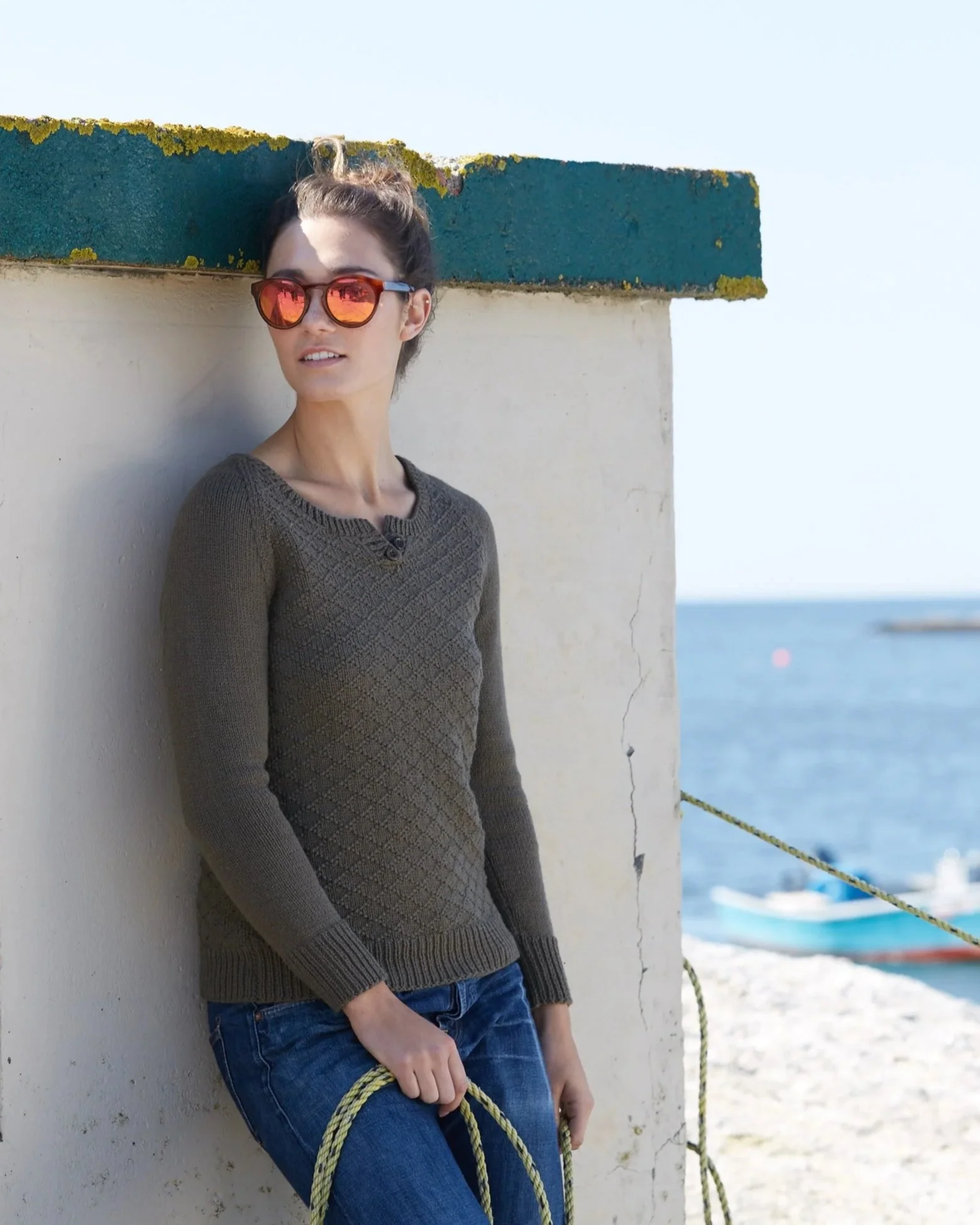A woman with dark hair tied up, wearing sunglasses, a patterned brown sweater, and blue jeans, leaning against a white wall near the ocean with boats in the background.