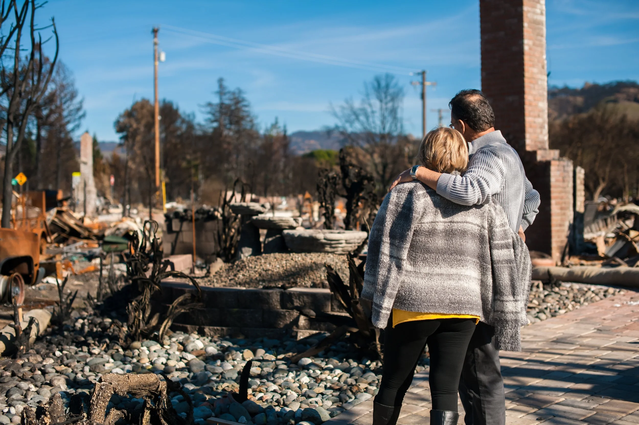 A man and a woman hugging each other outdoors among charred remains and debris, with burnt trees and a house chimney in the background.