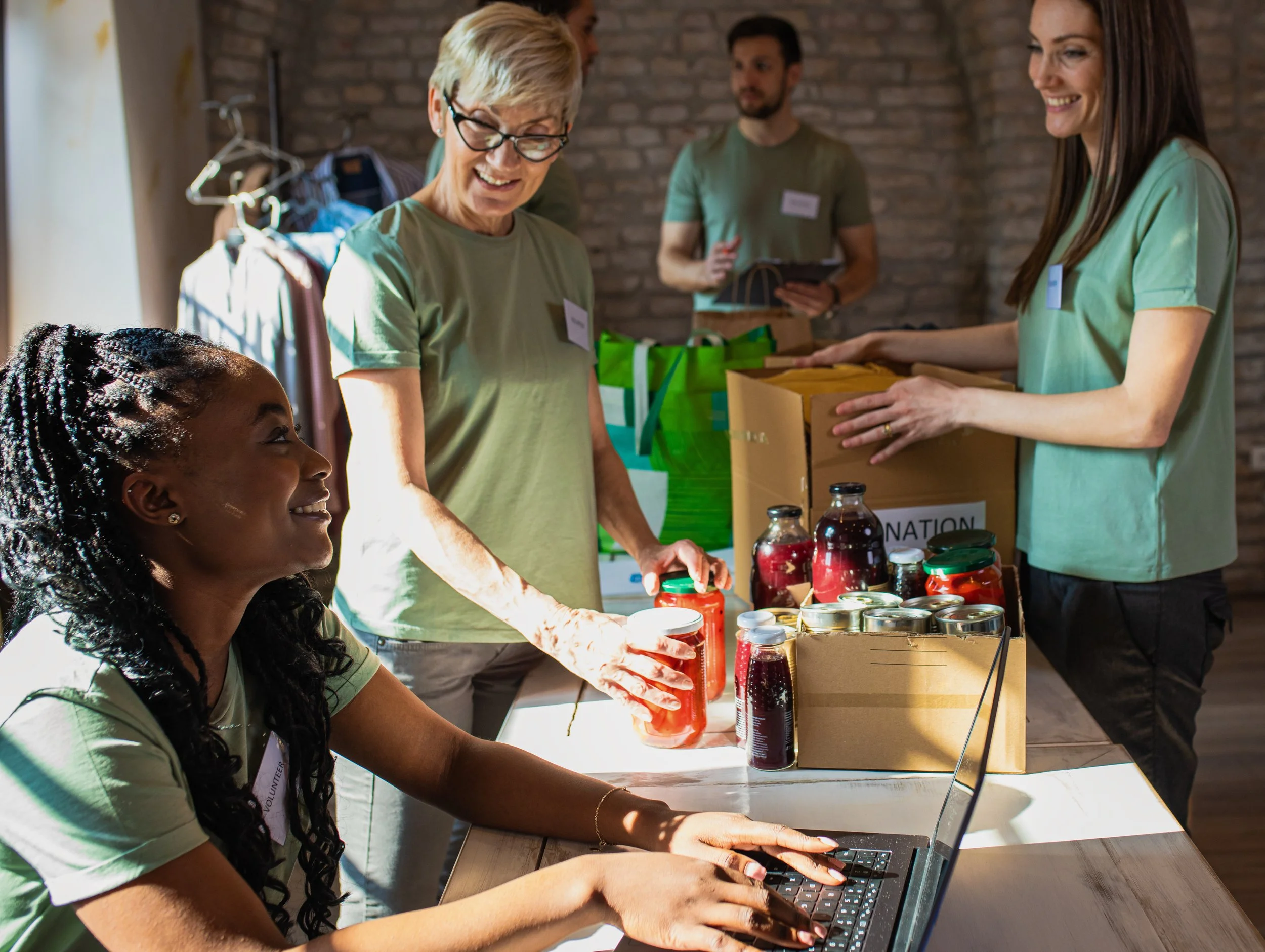 Several volunteers at a donation event, one woman with glasses and short blonde hair smiling and looking down at the table, another woman with long brown hair handing over a box, a girl with braided hair sitting at a laptop, and other volunteers in the background organizing items and paperwork.