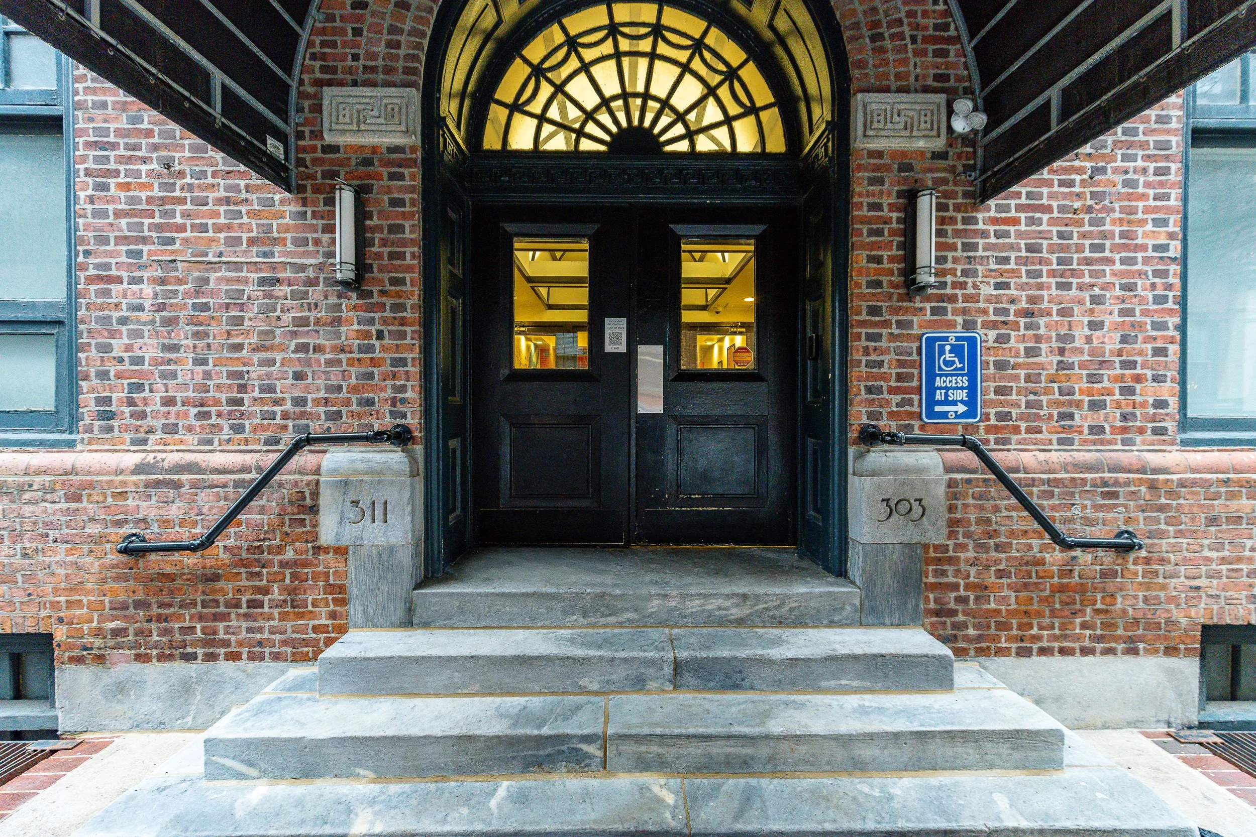 Front entrance of a brick building with black double doors, stone steps, and a sign indicating wheelchair access on the right side.