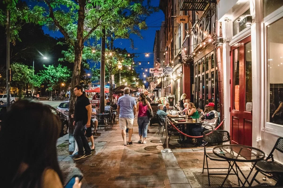 People walking and dining outside at night on a lively city sidewalk with string lights and storefronts.
