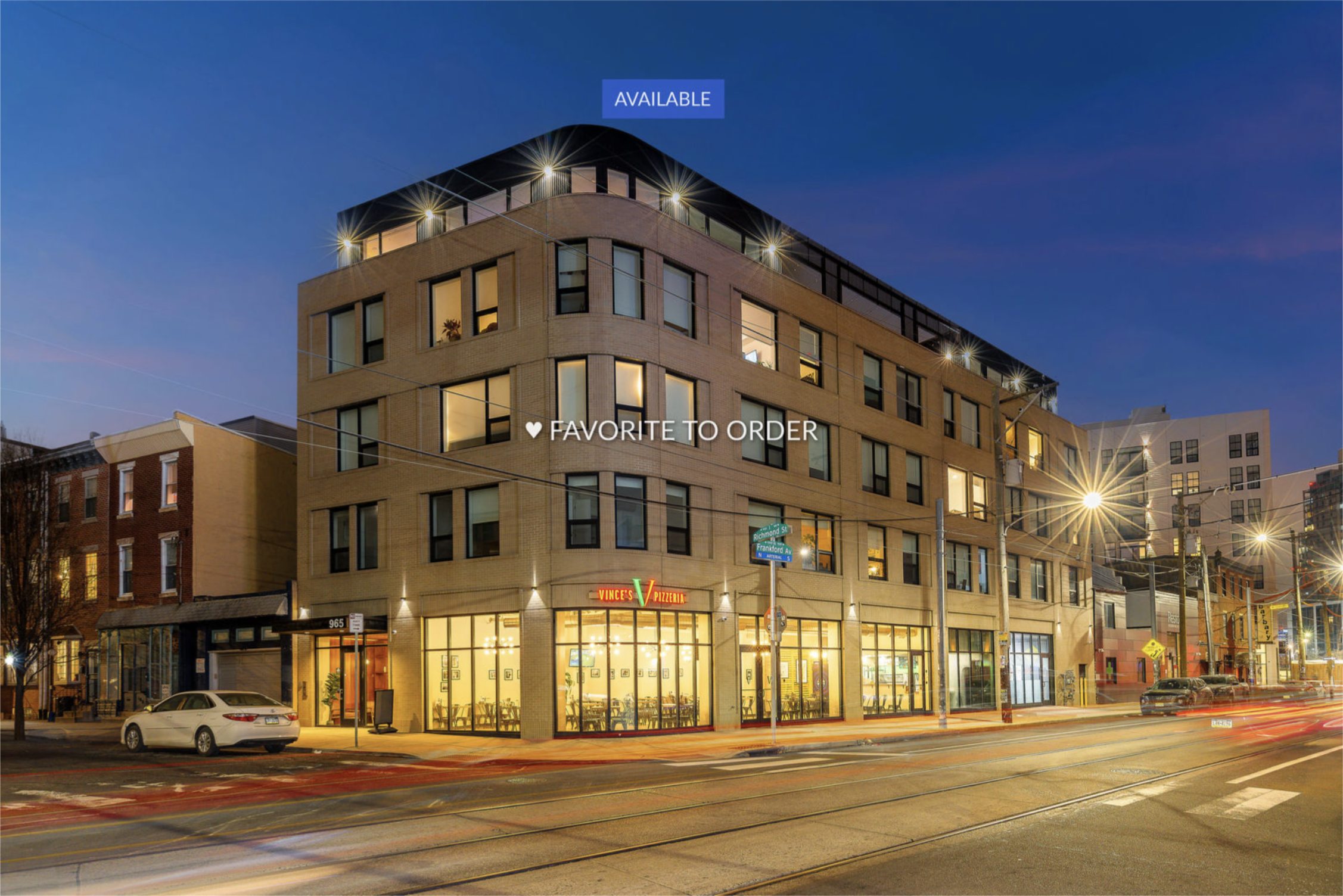 Street view of a modern multi-story building at dusk with illuminated interior and exterior lighting, a pizza restaurant on the ground floor, parked cars, and streetlights in front.