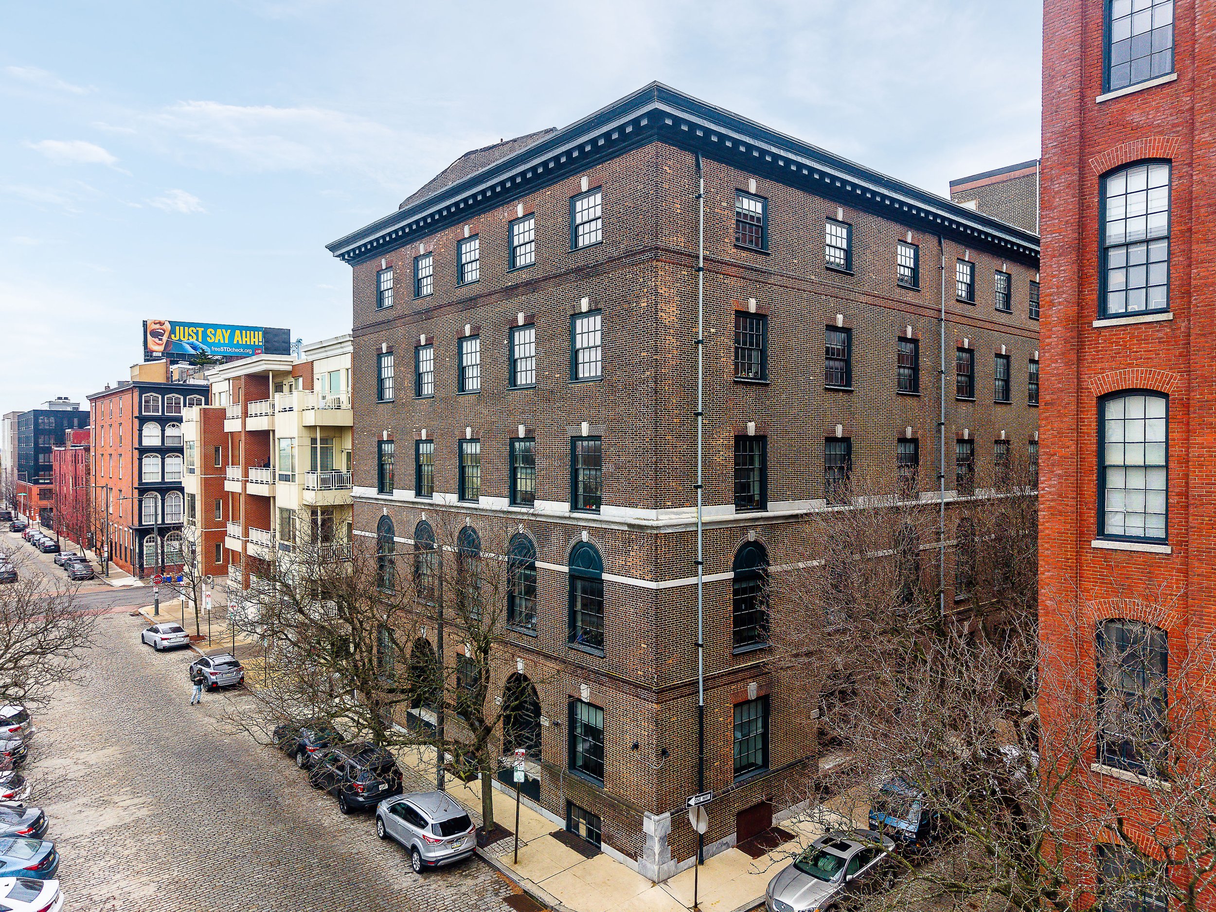A city street scene with a corner brick building with multiple windows, cars parked along the curb, leafless trees, and a billboard in the background that says "JUST SAY AH!"