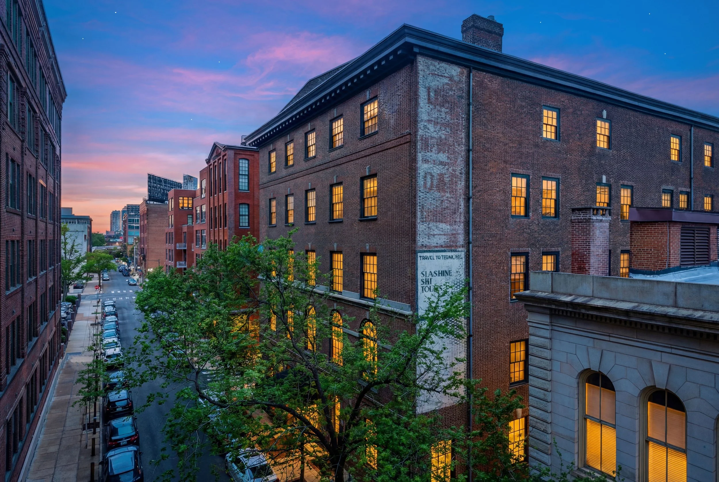 City street with brick buildings, trees, parked cars, and a sunset sky with pink and purple clouds.