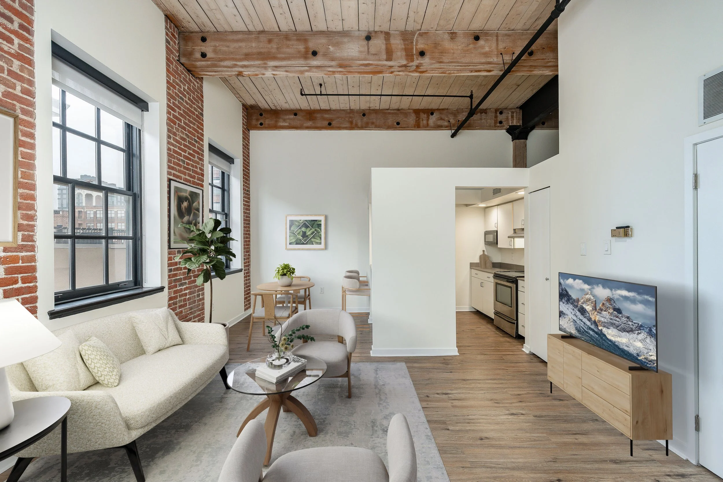 Living room with large windows, brick and white walls, wooden ceiling beams, furniture including a sofa, chairs, a round coffee table with books, and a television displaying mountain scenery.