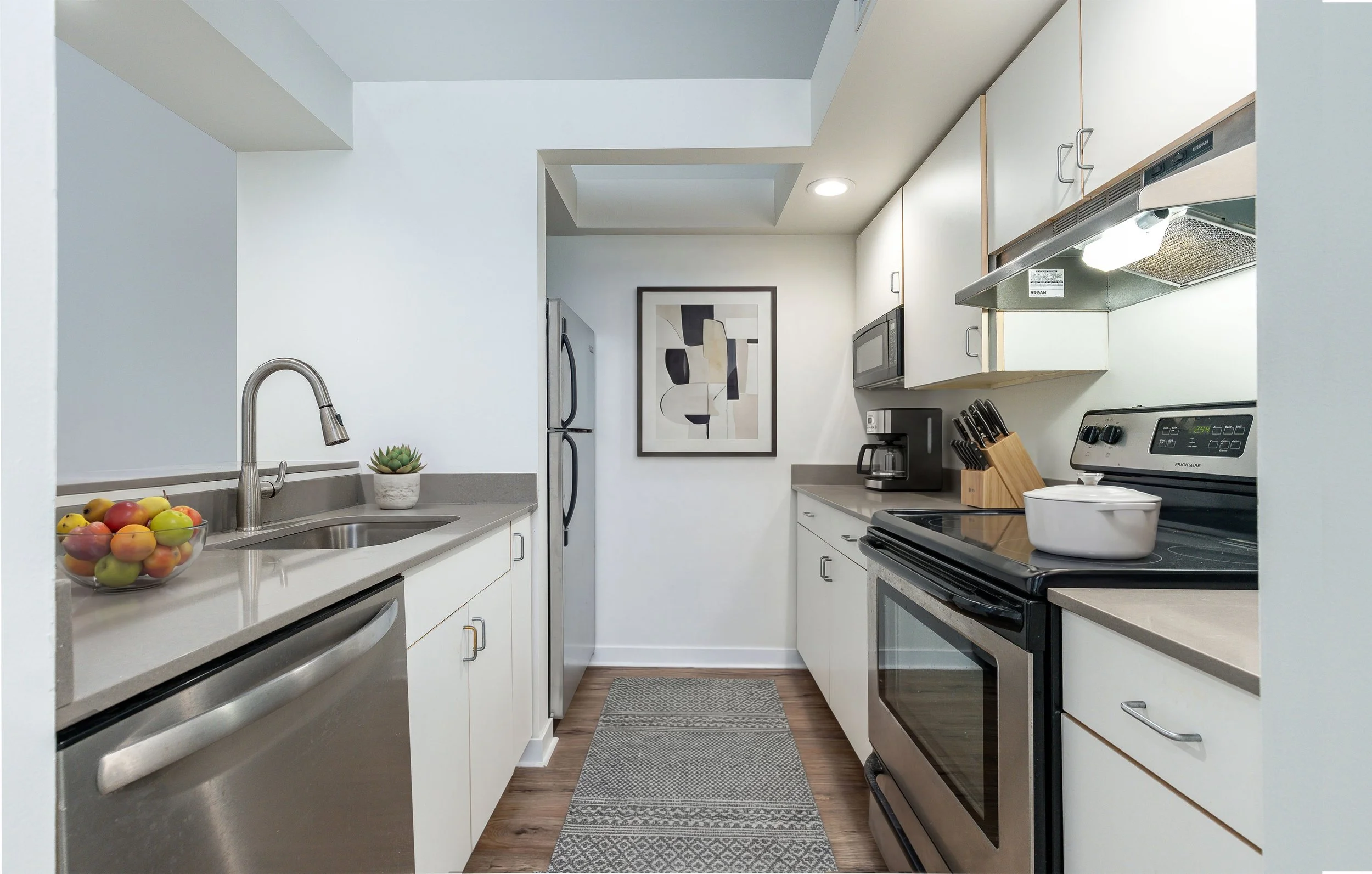 A compact, modern kitchen with white cabinets and gray countertops. Features include a stainless steel sink with a faucet, a bowl of apples and peaches, a small potted plant, a coffee maker, a knife block, a microwave, and a stove with an oven. There is a framed abstract art piece on the wall, hardwood flooring, and built-in ceiling lighting.
