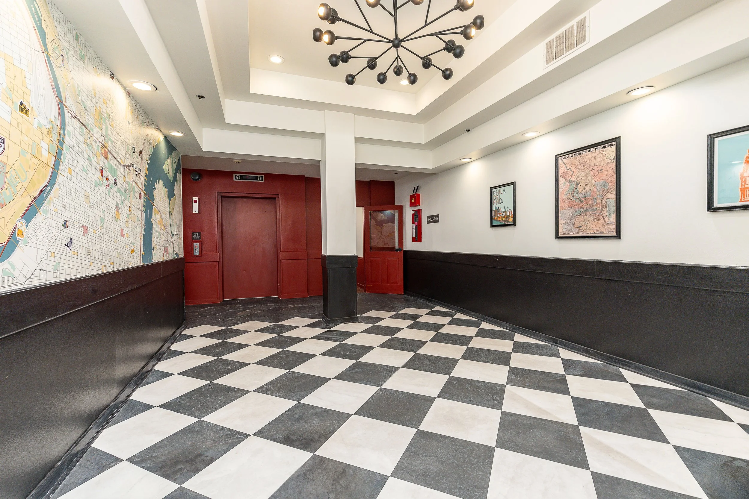 An indoor space with black and white checkered flooring, white walls with framed maps, red doors, and a black modern chandelier hanging from the ceiling.