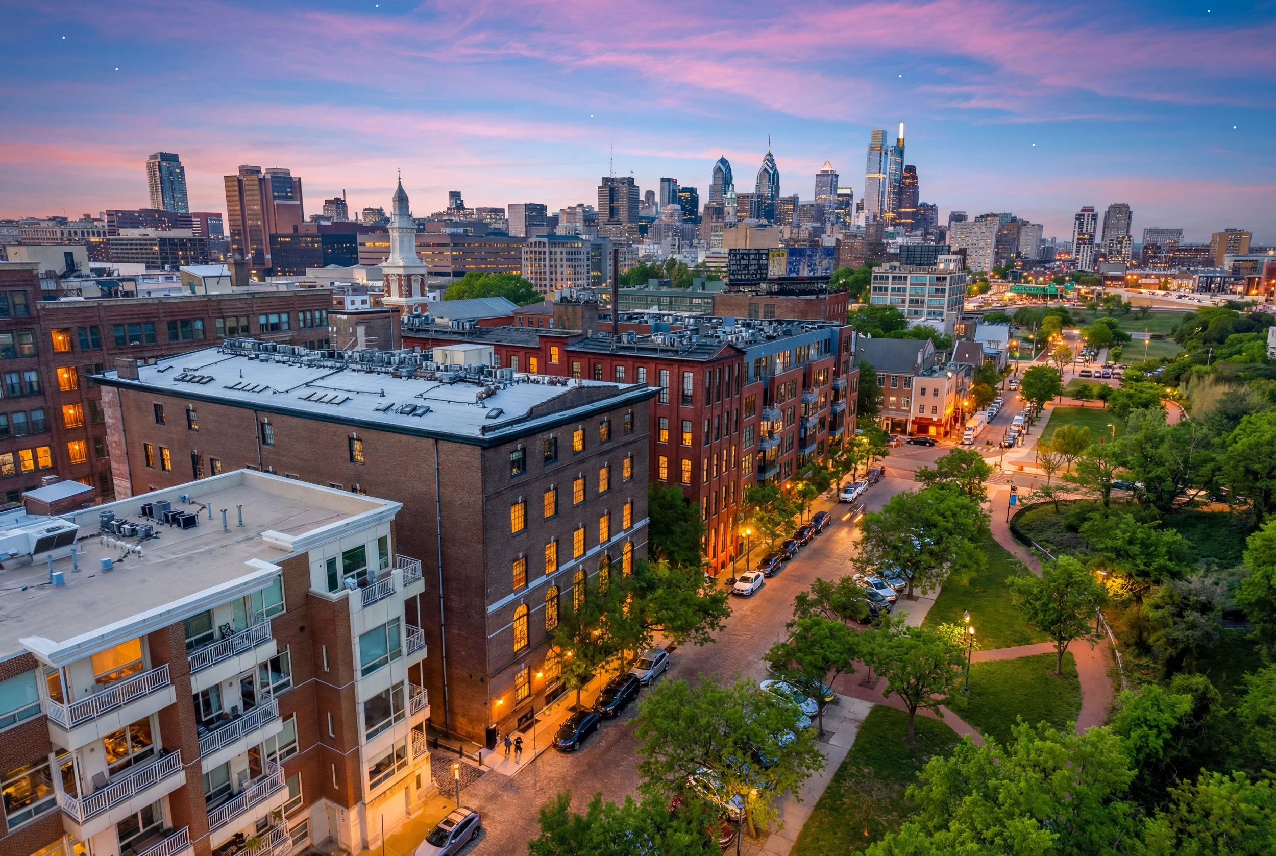 City skyline at dusk with tall buildings, some with illuminated windows, trees lining the streets, and a colorful sky with pink and purple clouds.