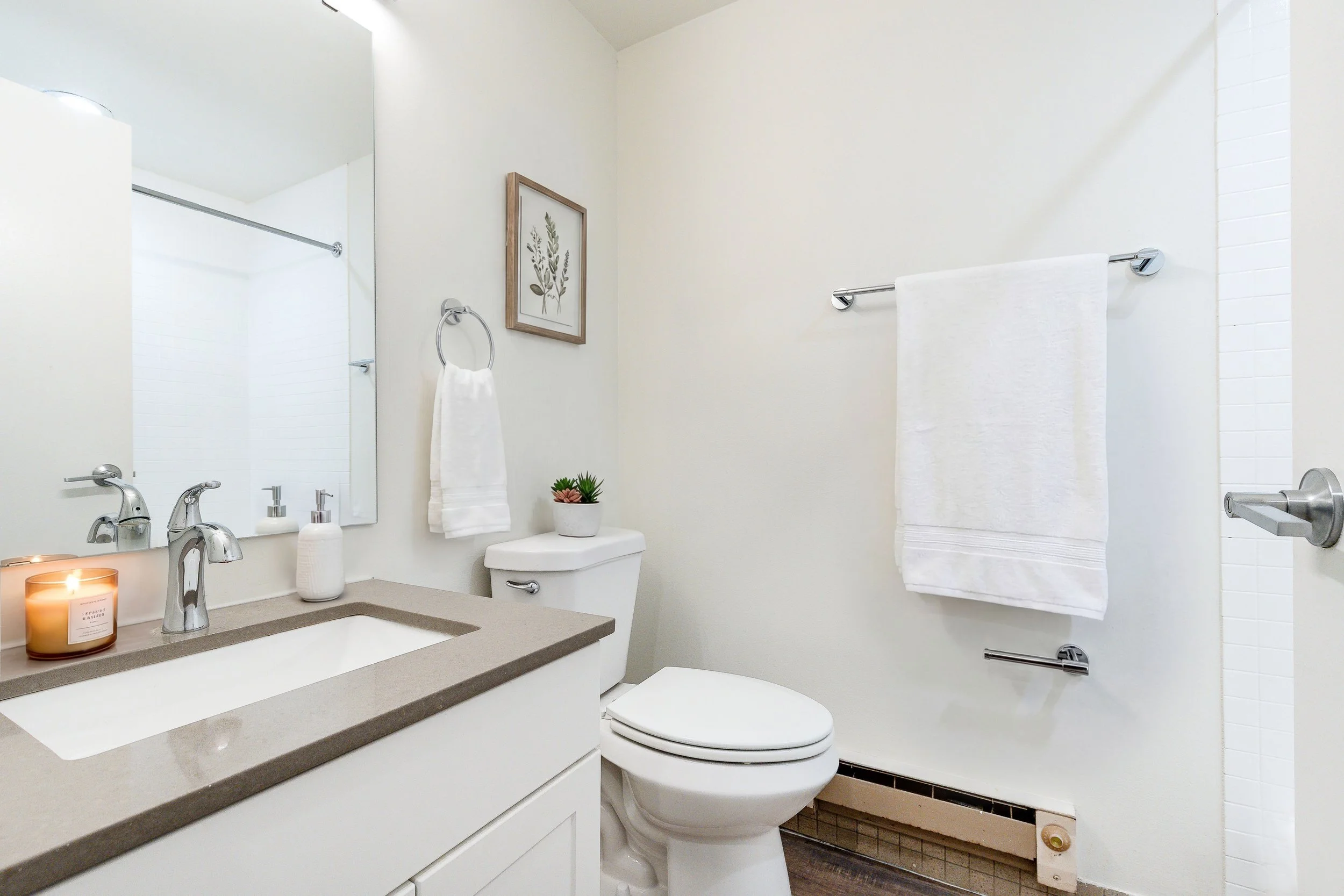 A modern bathroom featuring a white vanity with a gray countertop, a sink, and chrome faucet. There are two soap dispensers, a lit candle, and a small potted plant on the vanity. A large mirror is mounted above it. The toilet is situated next to the 