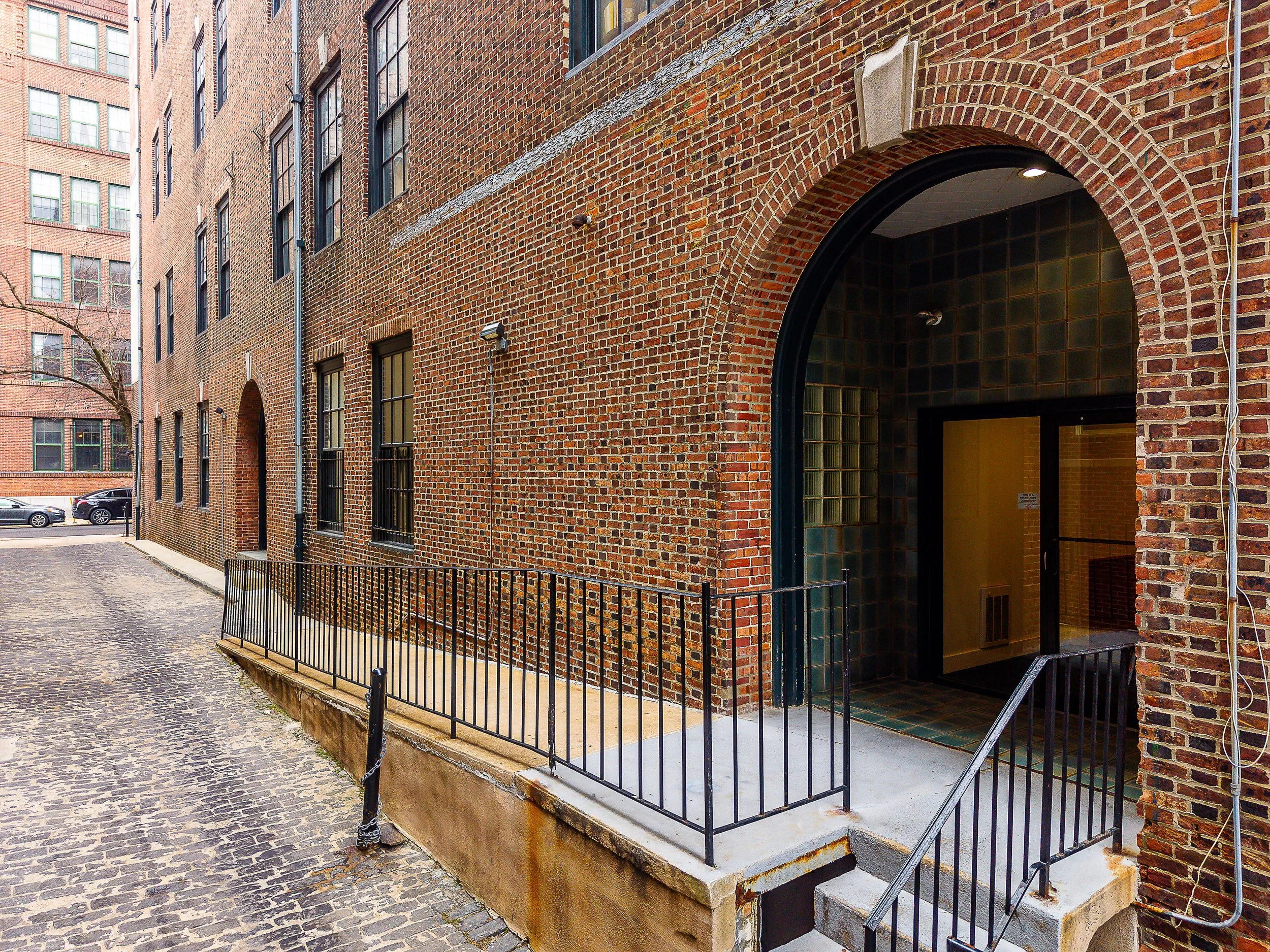 Brick apartment building with arched entrance and metal railing stairs leading up to the door, cobblestone sidewalk, parked cars, and leafless trees in the background.