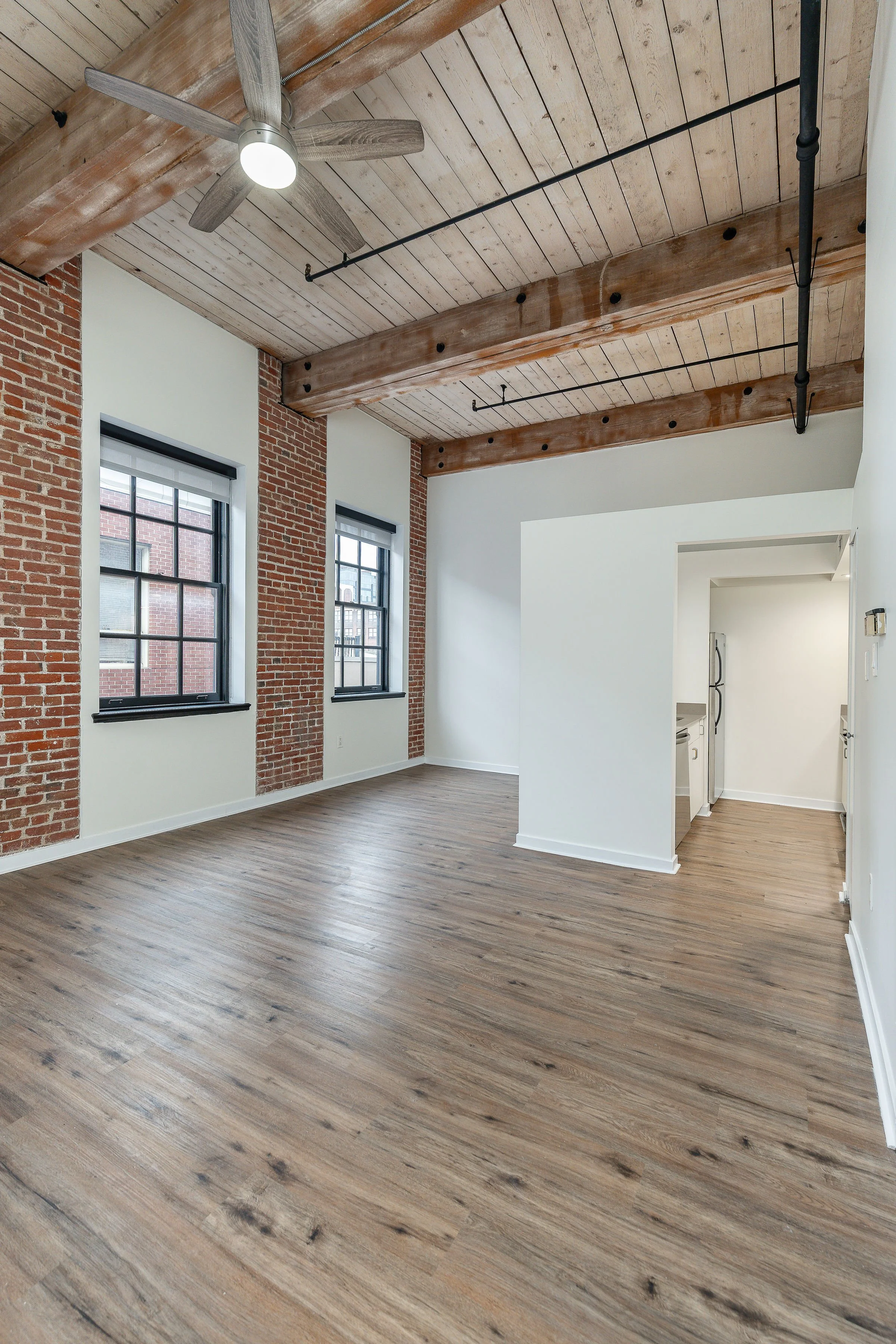 Bright, open loft apartment with exposed brick walls, large windows, wood plank ceilings, modern ceiling fan, and wooden flooring.