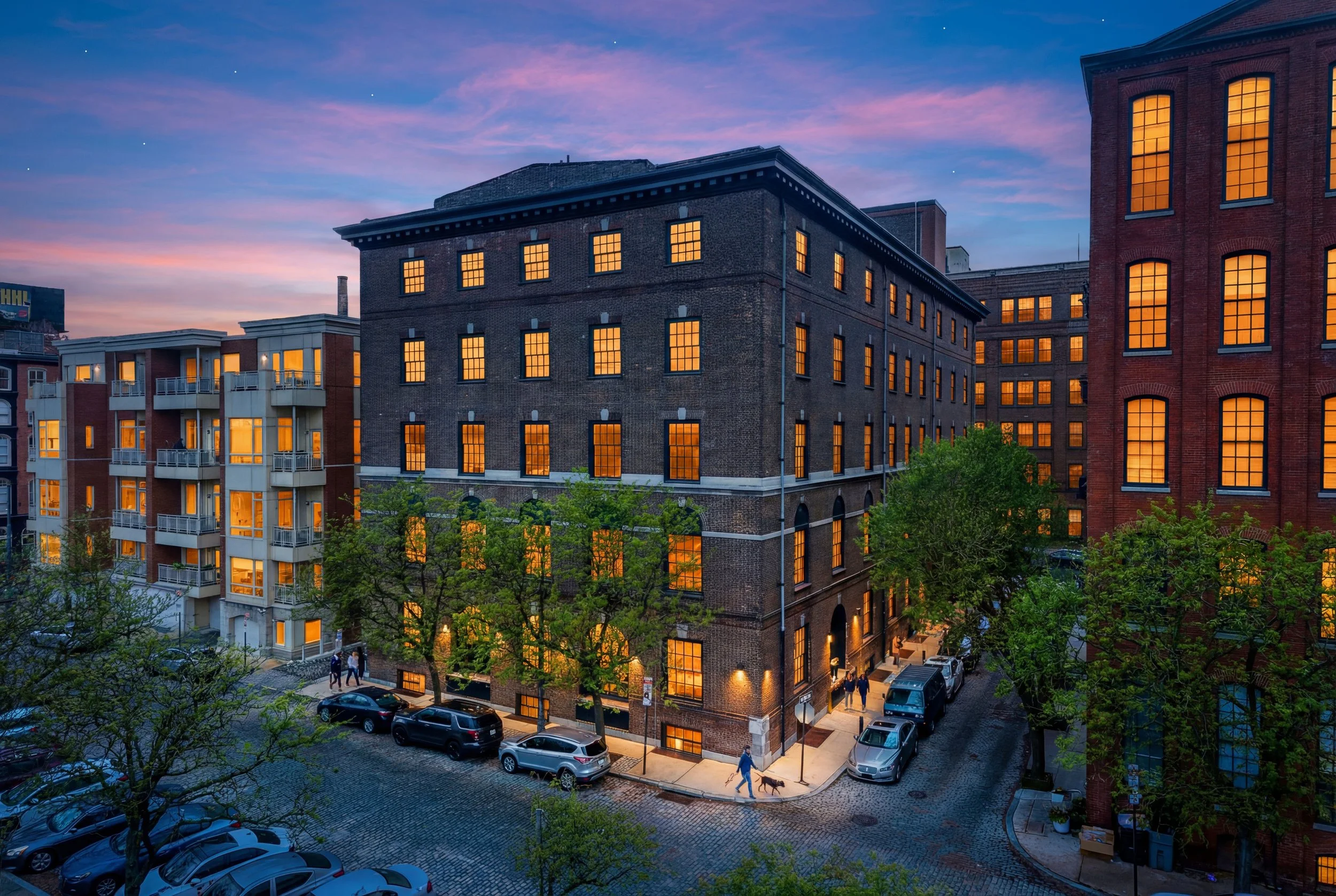 City street scene at dusk with buildings, people walking dogs, parked cars, trees, and illuminated windows.