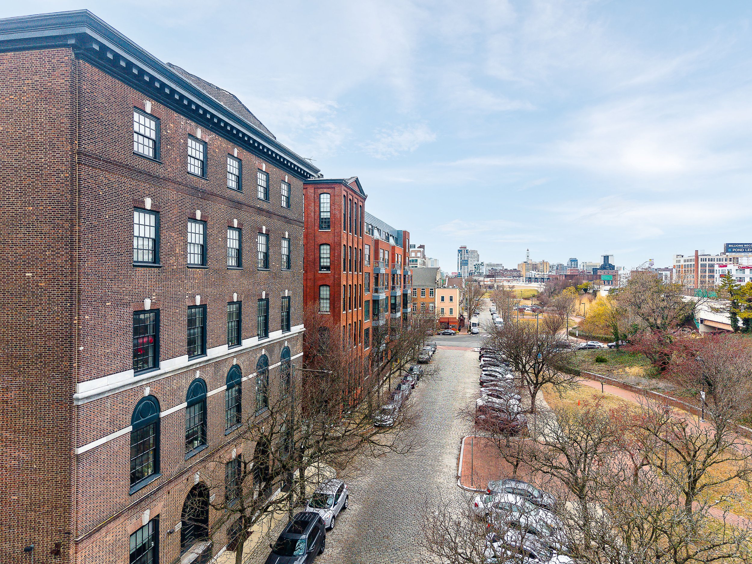 A cityscape view from an elevated perspective showing a row of brick residential buildings with multiple windows, a cobblestone parking area with several parked cars, leafless trees, and a distant skyline with taller modern buildings under a partly c