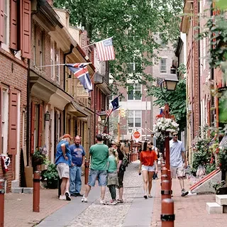 A lively city street with people walking and talking, surrounded by colorful buildings, with flags hanging across the street and flowers decorating the storefronts.
