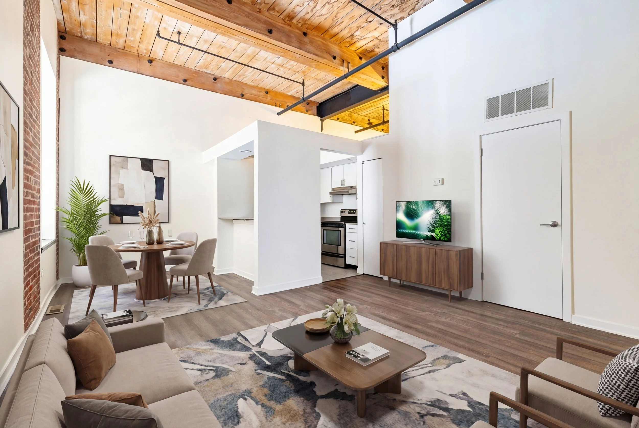 Open-concept living room with a brick wall, wooden ceiling beams, a beige sofa with pillows, a wooden coffee table, a patterned rug, a dining area with a round table and upholstered chairs, a potted plant, and a wall-mounted TV in the background.