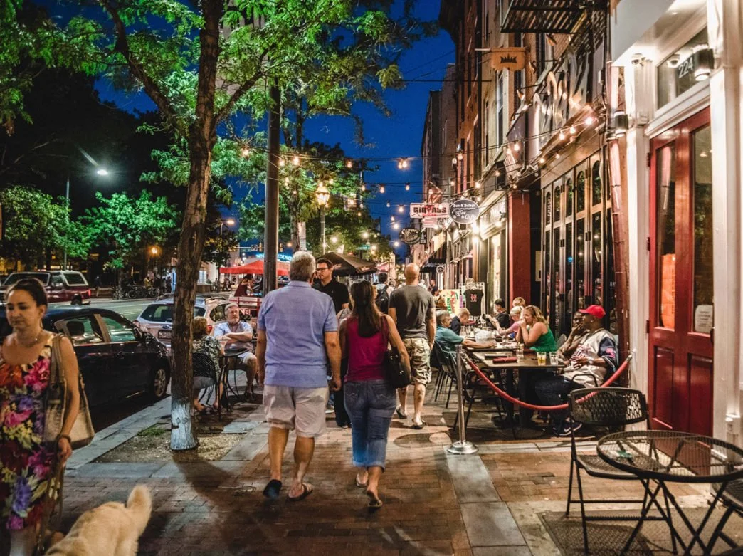 Nighttime street scene with people dining at outdoor tables outside a restaurant, illuminated string lights overhead, a tree, parked cars, and urban buildings lining the sidewalk.
