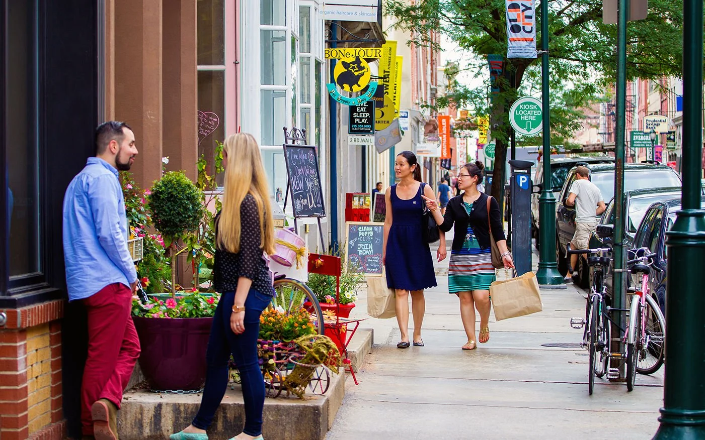 People talking on a busy city sidewalk with storefronts, colorful signs, bicycles, and trees.