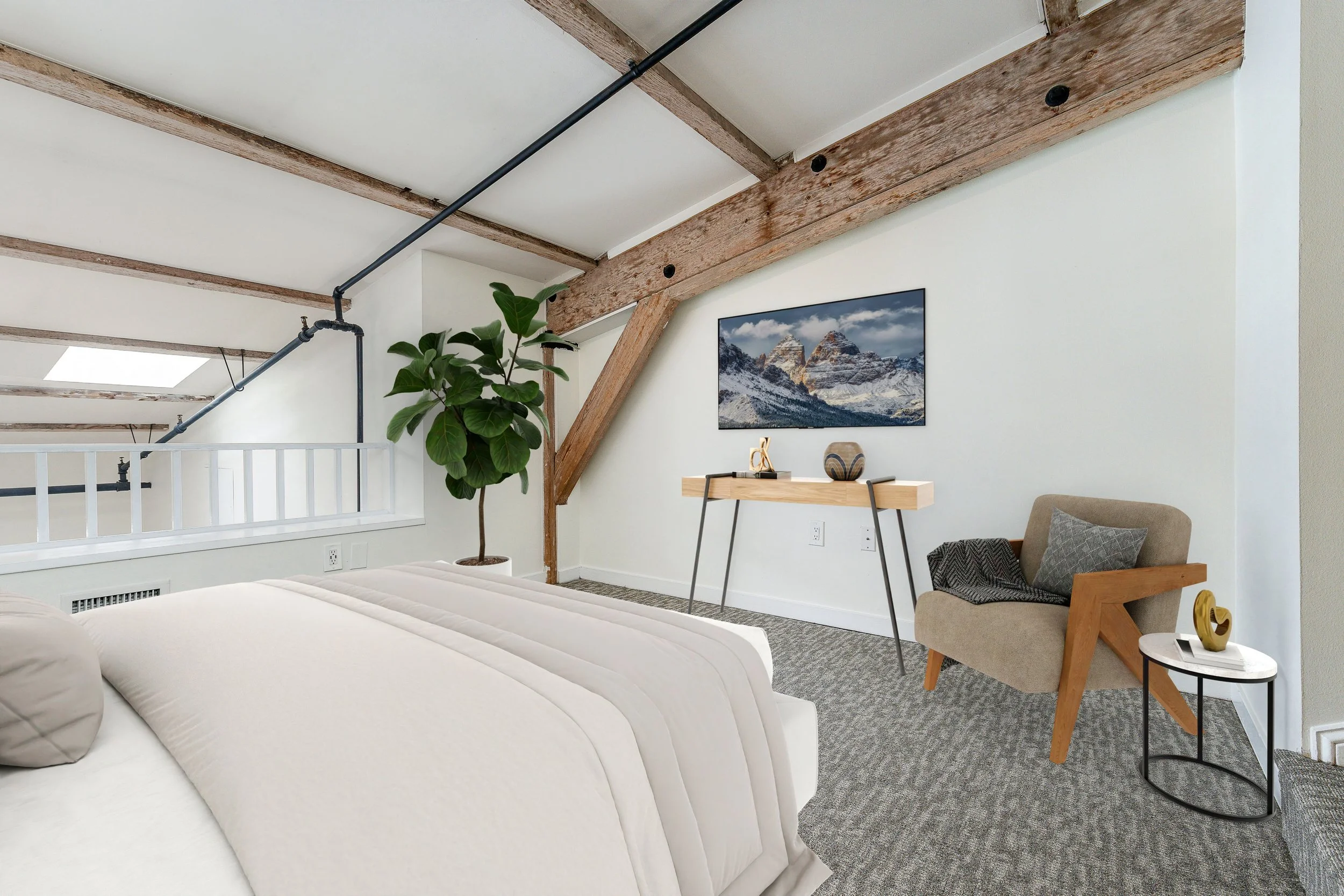 A cozy bedroom with beige bedding, a large green plant in a white pot, a wooden desk with decorative items, a framed mountain landscape photograph, a beige armchair with gray pillows, and exposed wooden beams on the ceiling. There is a skylight window and a white railing near the bed.