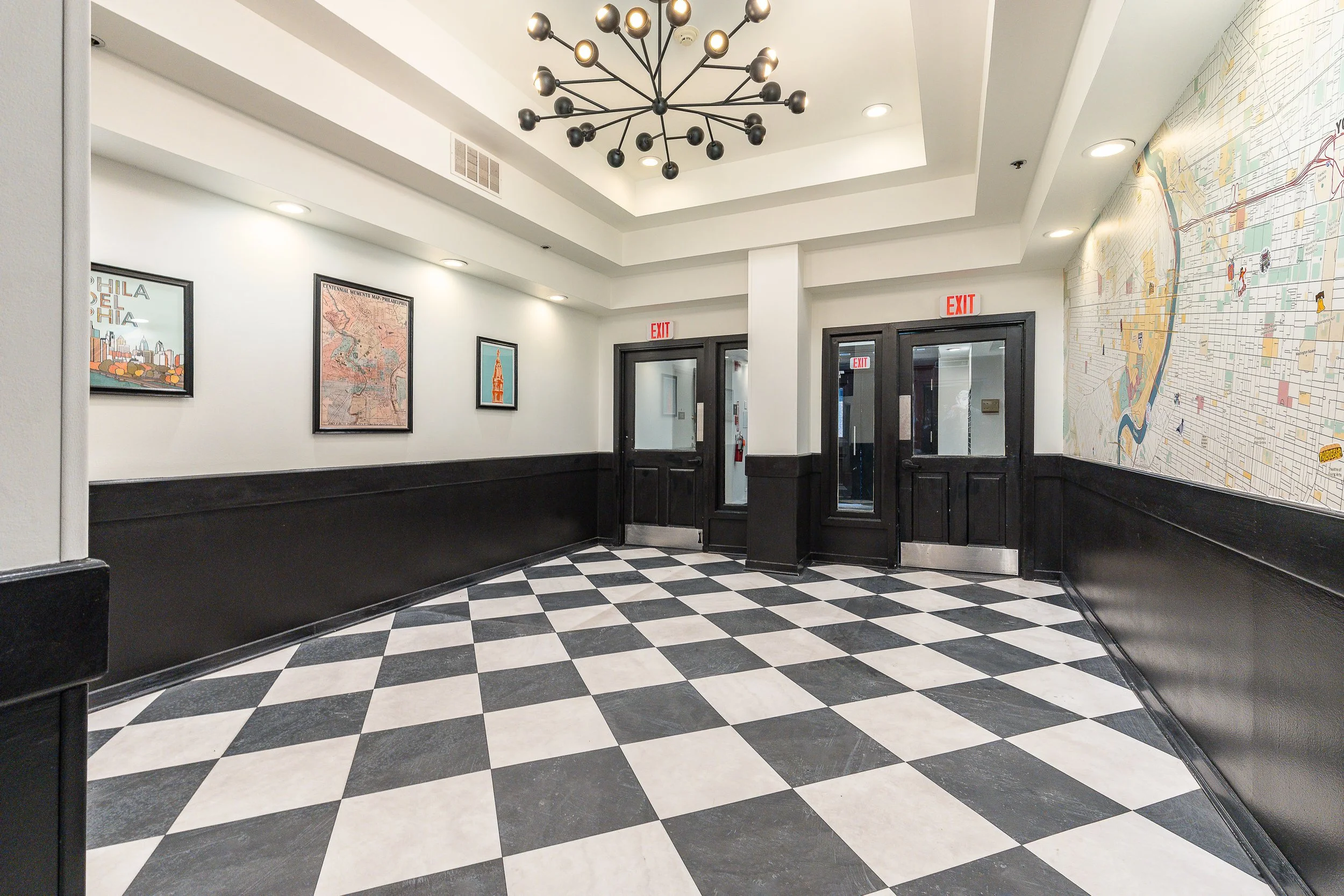 An interior hallway with black and white checkered flooring, white walls, framed artwork, two exit doors, and a large modern chandelier ceiling light.