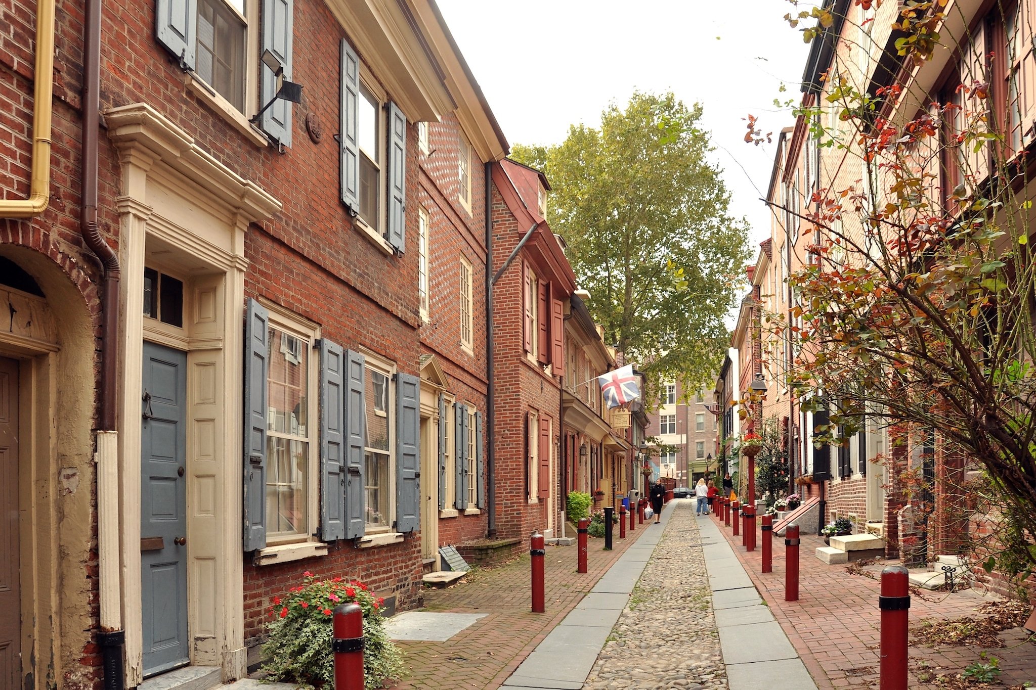 A narrow sidewalk with cobblestone and concrete sections, lined with red metal bollards, alongside row houses with brick facades and colorful shutters, trees with green and some reddish leaves, and a Union Jack flag hanging on one building.