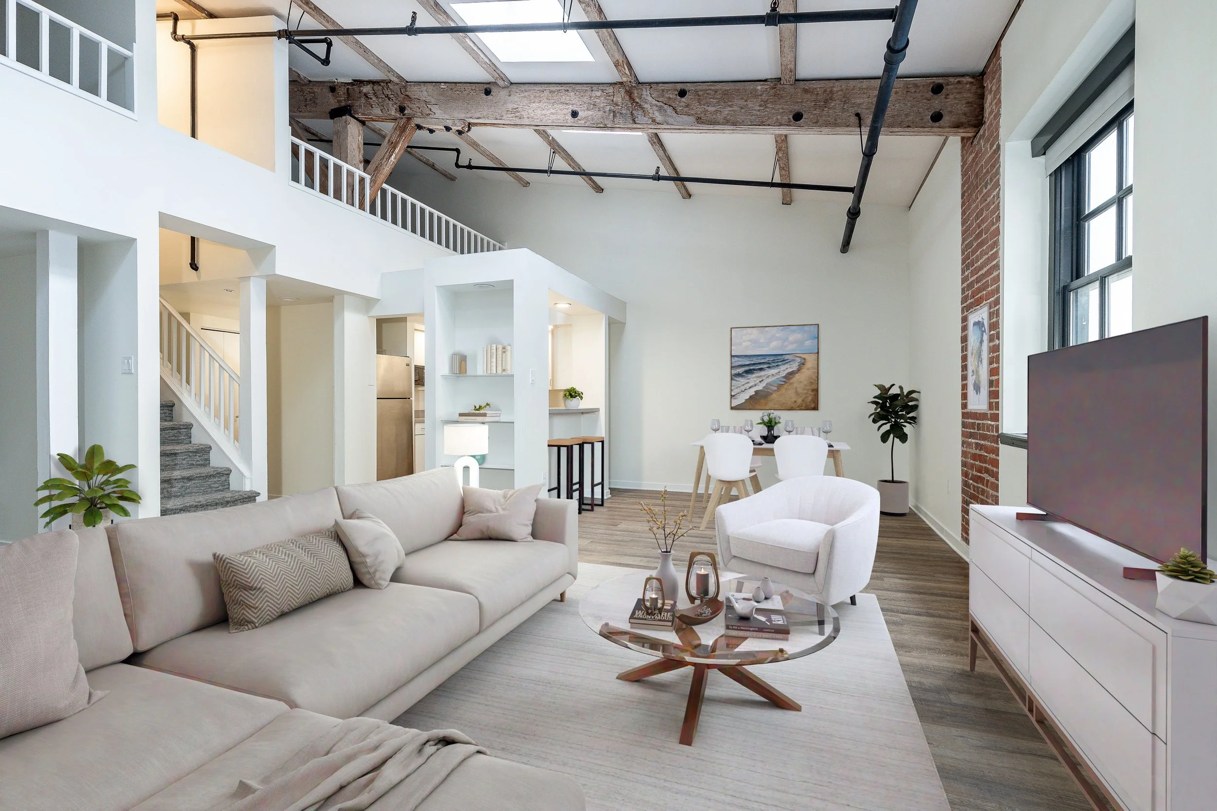Modern living room interior of The Tannery in Old City Philadelphia. A white sofa, armchair, wooden coffee table with decor items, potted plants, wall art, large windows, exposed brick wall, and open ceiling with wooden beams and black pipes.