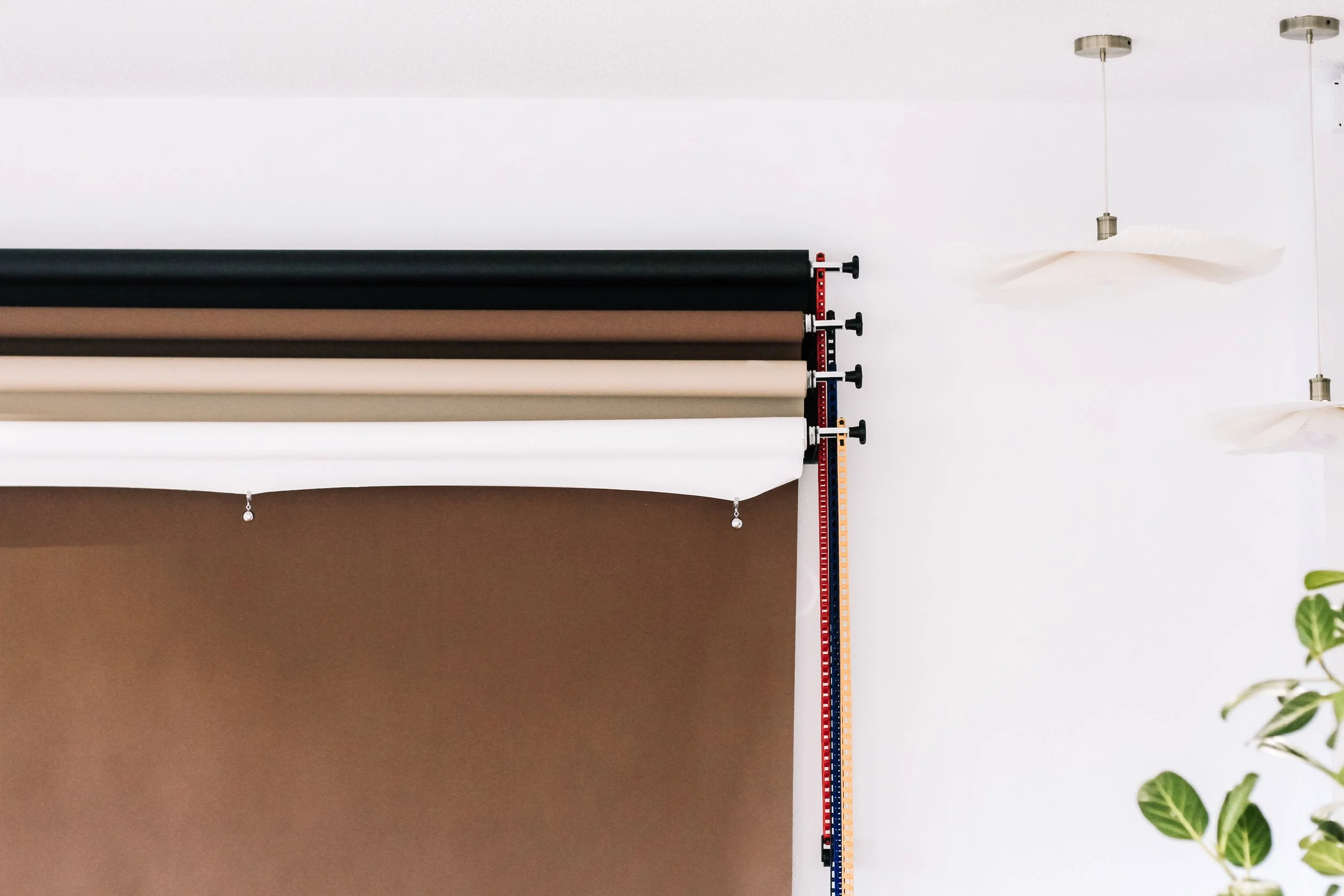 Display of fabric rolls in black, dark brown, and light brown colors mounted on a wall with a measuring ruler and a modern hanging light fixture also visible.