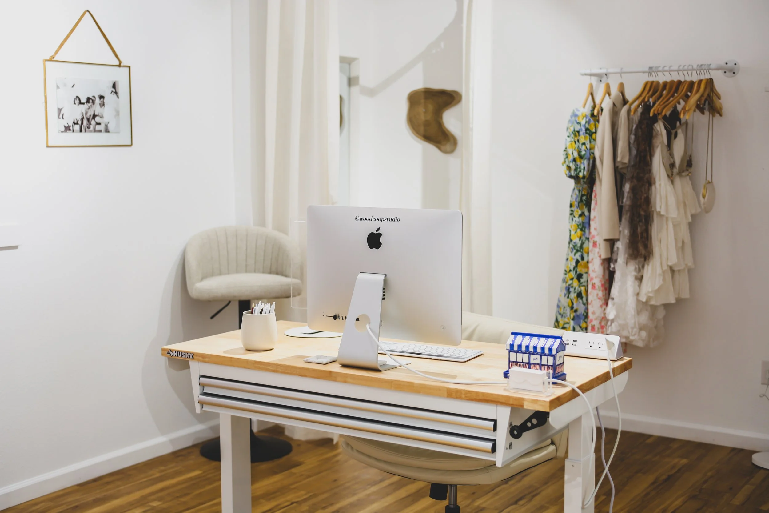 A medical or consultation room with a white wall, a light-colored wooden desk, and a white computer monitor. There is a beige upholstered chair behind the desk, and a white shelf with a few small items on the desk. On the right, a white clothing rack with various dresses and fabrics hangs. In the background, a mirror, a decorative wooden wall piece, and a framed black-and-white photograph of children are visible.