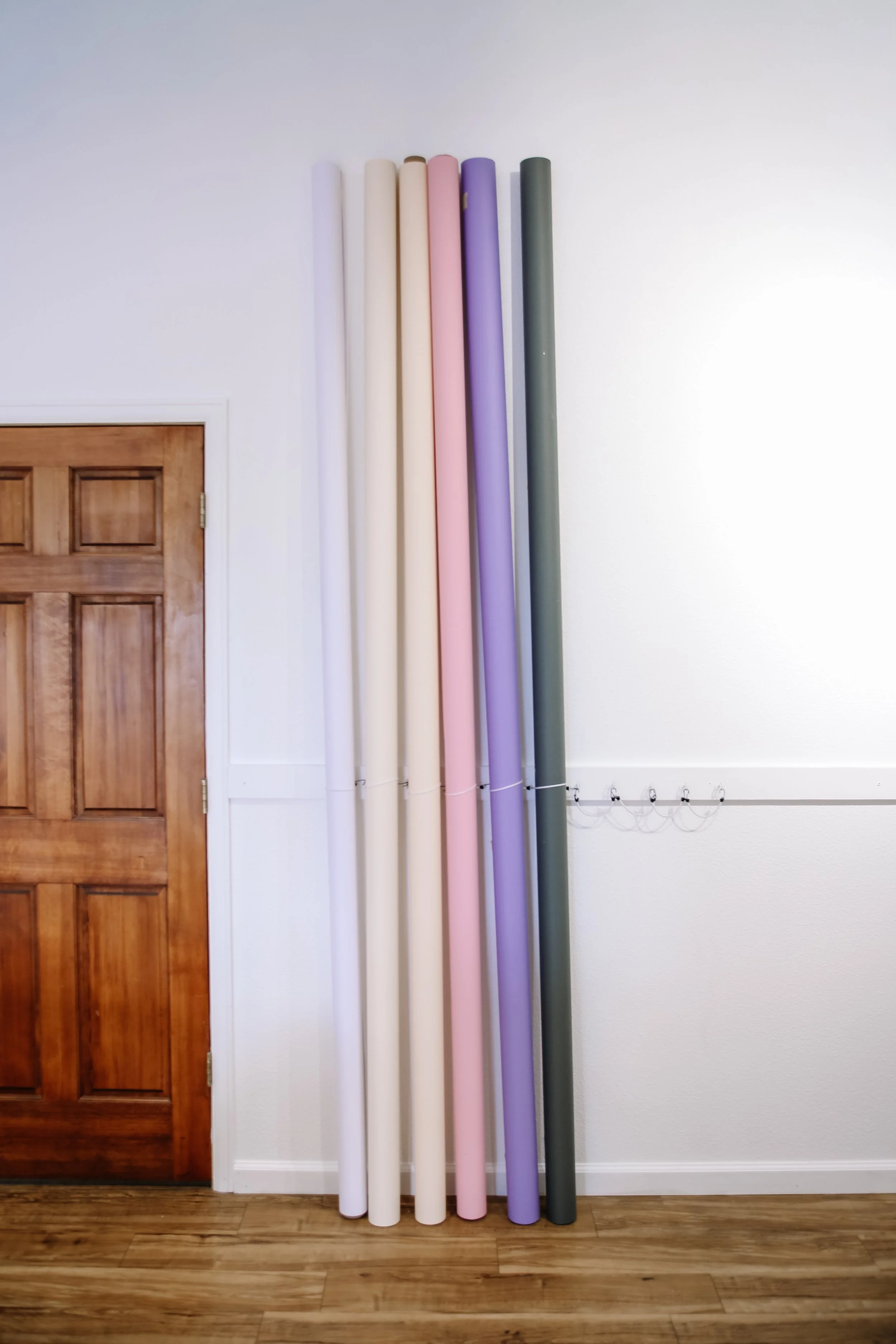 Seven tall, colorful pipes or poles standing vertically beside a wooden door and white wall with some electrical wiring at the bottom.