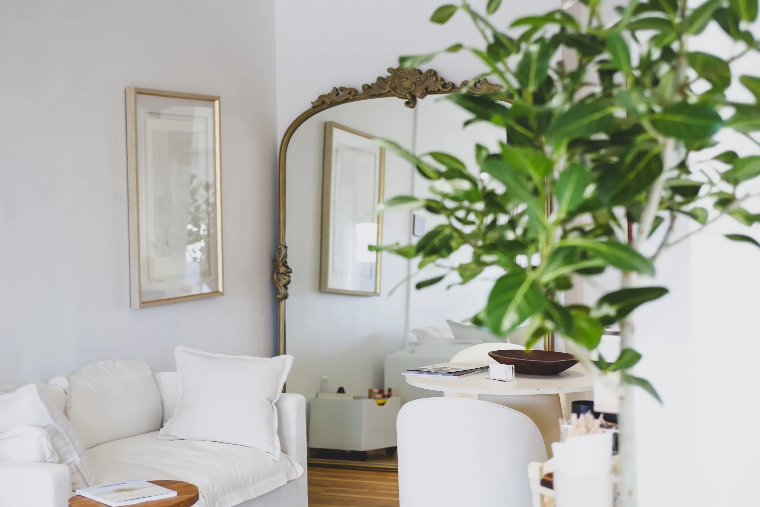 A cozy living room with white walls, a white sofa with white pillows, and a small wooden table with books. A large green houseplant is in the foreground. On the wall are two framed pictures and an ornate mirror. There is a white round table with some