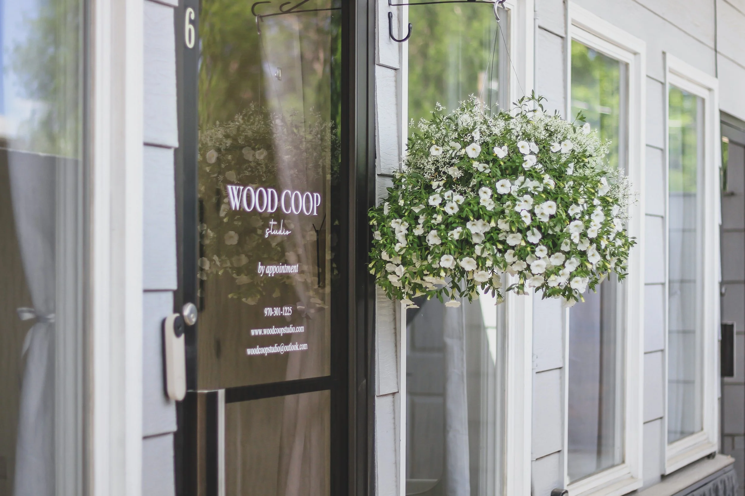 The entrance door of Wood Co-op studio, with a glass panel displaying the studio name, contact information, and email, surrounded by white siding, and a hanging flower basket filled with white petunias.