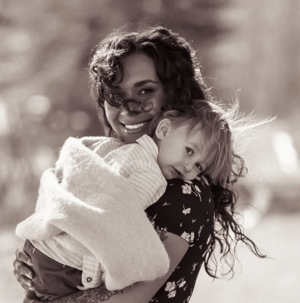 A woman with curly hair smiling and holding a young girl with straight hair in a close embrace outdoors.