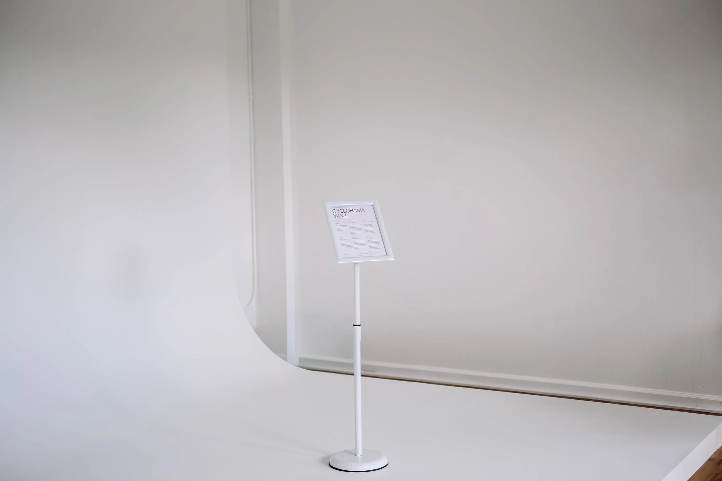 A white floor lamp with a small white sign displaying information, standing against a plain white wall.