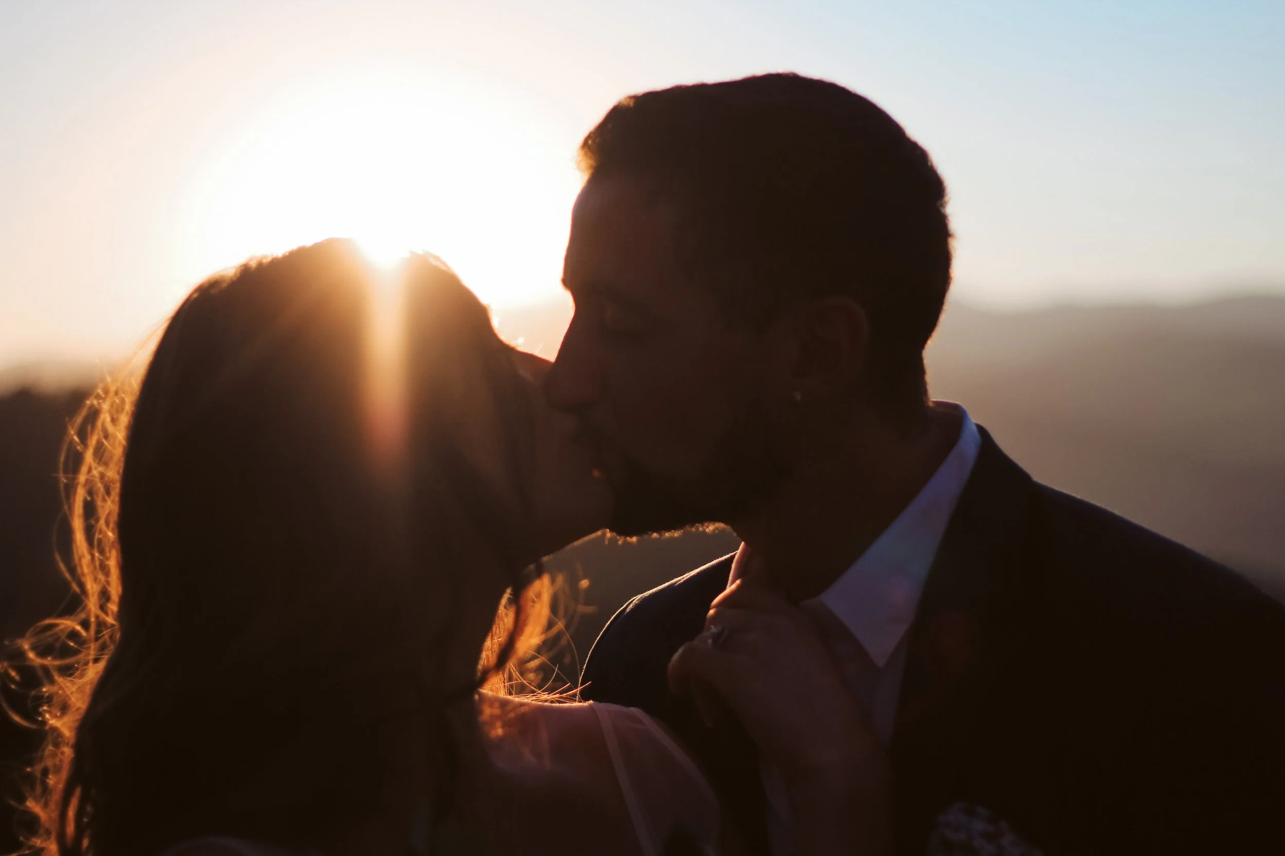A couple sharing a kiss at sunset, with the sun setting behind them and the landscape in the background.