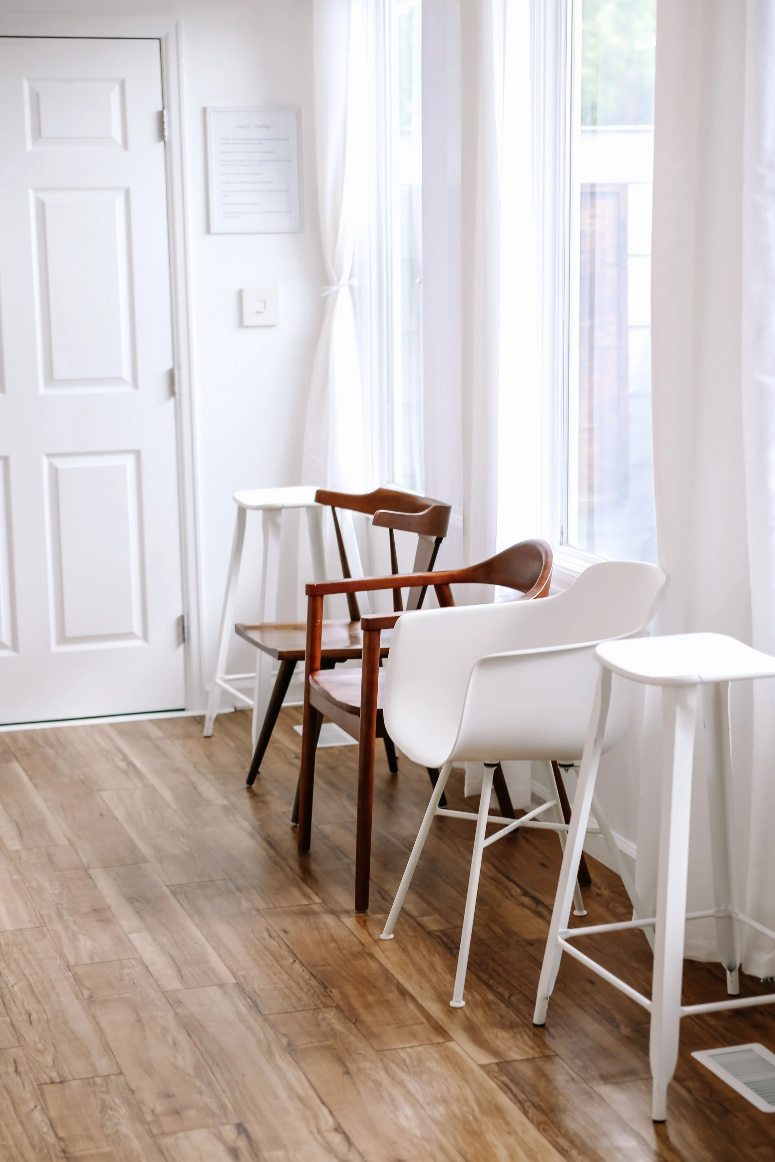 A row of modern chairs in various colors and styles lined up against a white wall with large windows, sheer curtains, and a framed picture on the wall in a bright, minimalistic room with wooden flooring.