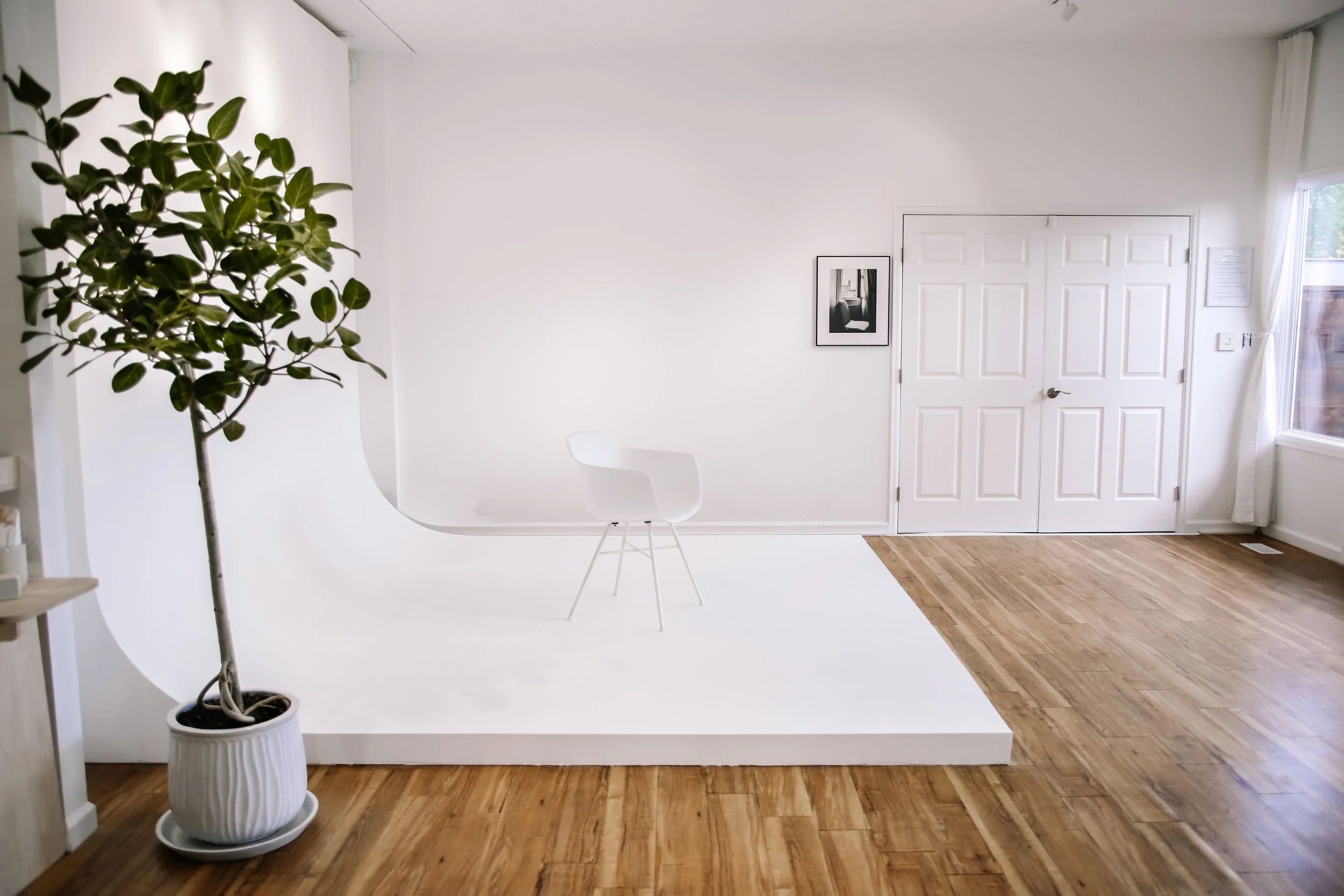 A minimalist interior room with white walls and a hardwood floor. There is a large houseplant in a white pot on the left, a white chair in the center, a black-and-white framed art print on the wall, and a closed white double door. A window with sheer